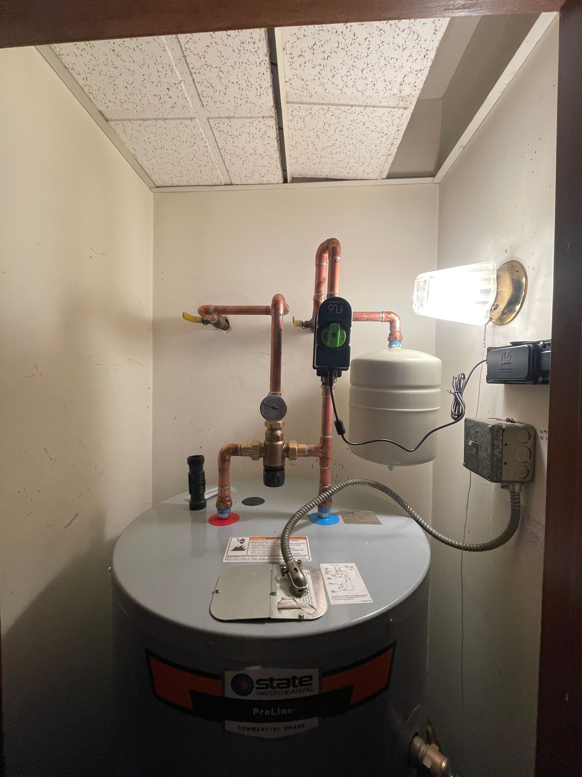 Water heater and plumbing in a small utility closet with beige walls and a drop ceiling.