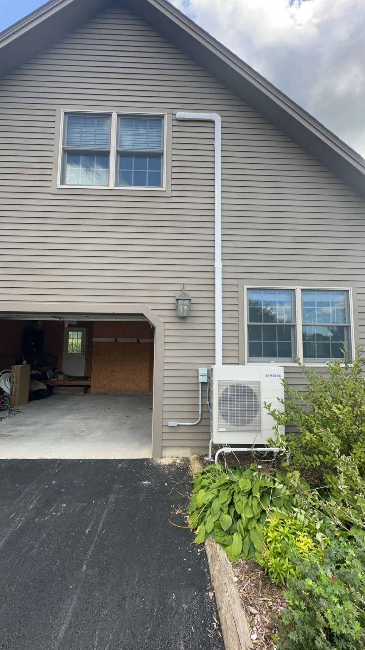 Two-story beige house with open garage, driveway, and white outdoor AC unit beside the front wall.