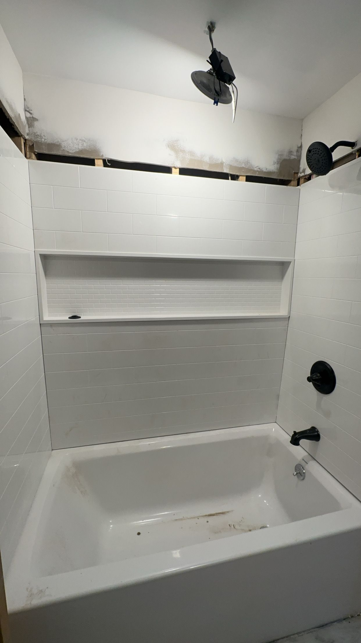 Partially tiled bathroom wall with a bathtub, shelf niche, and exposed fixtures under renovation