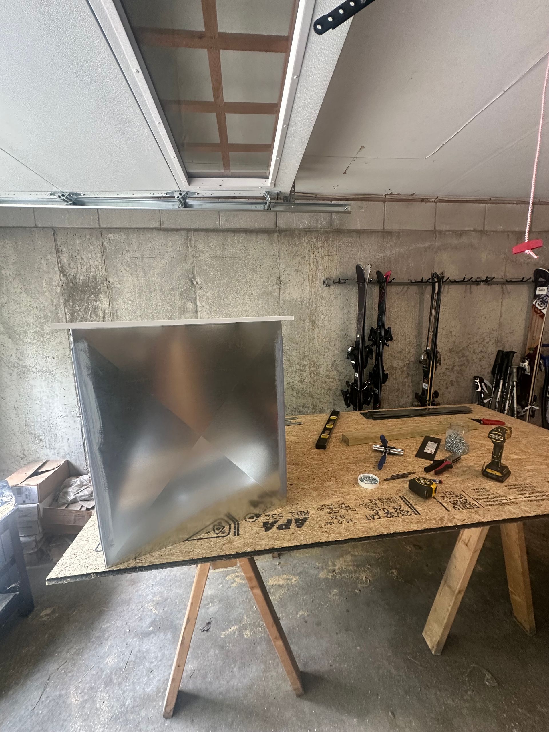 Workshop table with tools and a large metal panel leaning on the left under a slanted ceiling