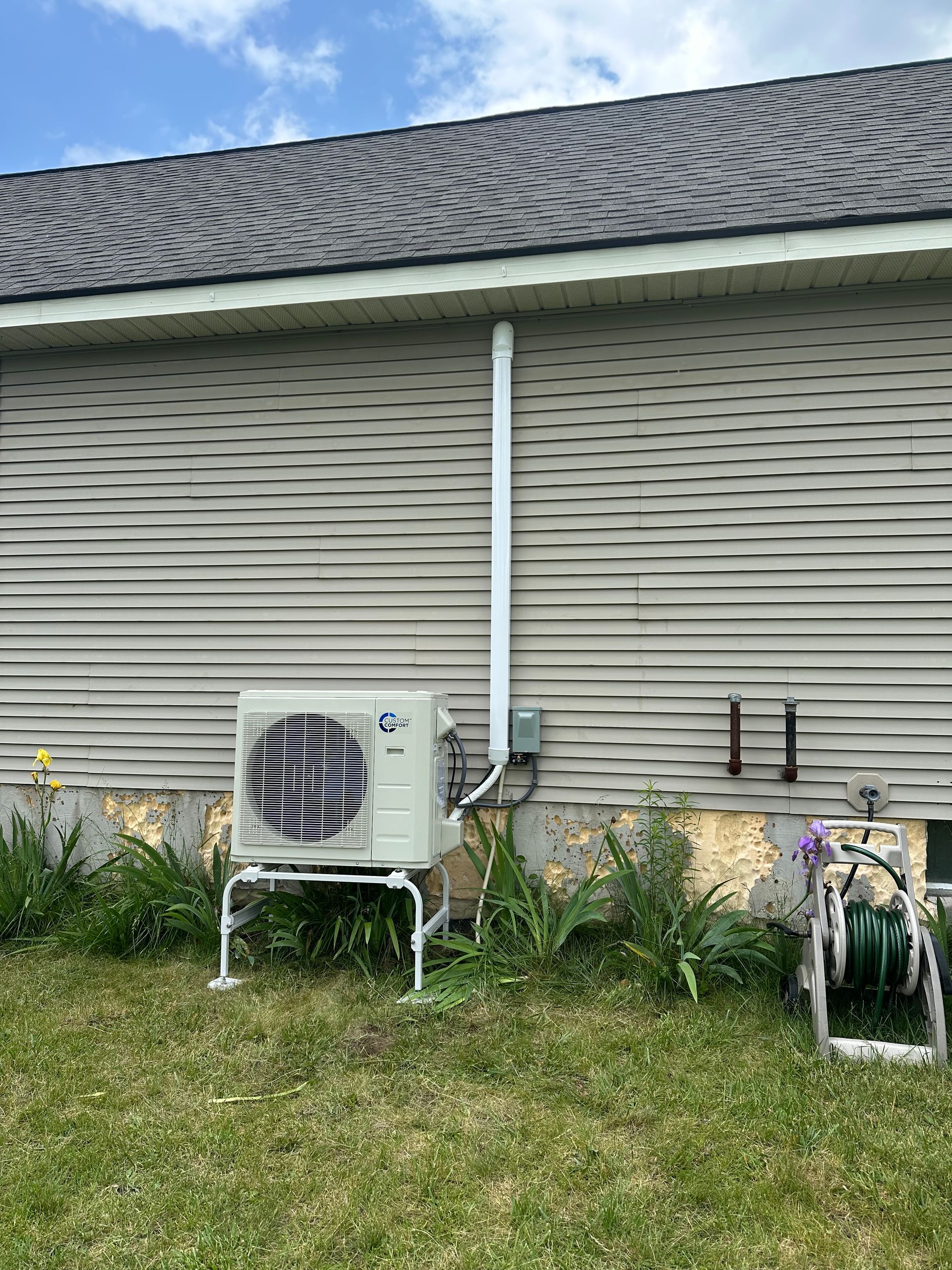 House siding with an outdoor AC unit, white conduit pipe, and grass in front of a shingled roof.