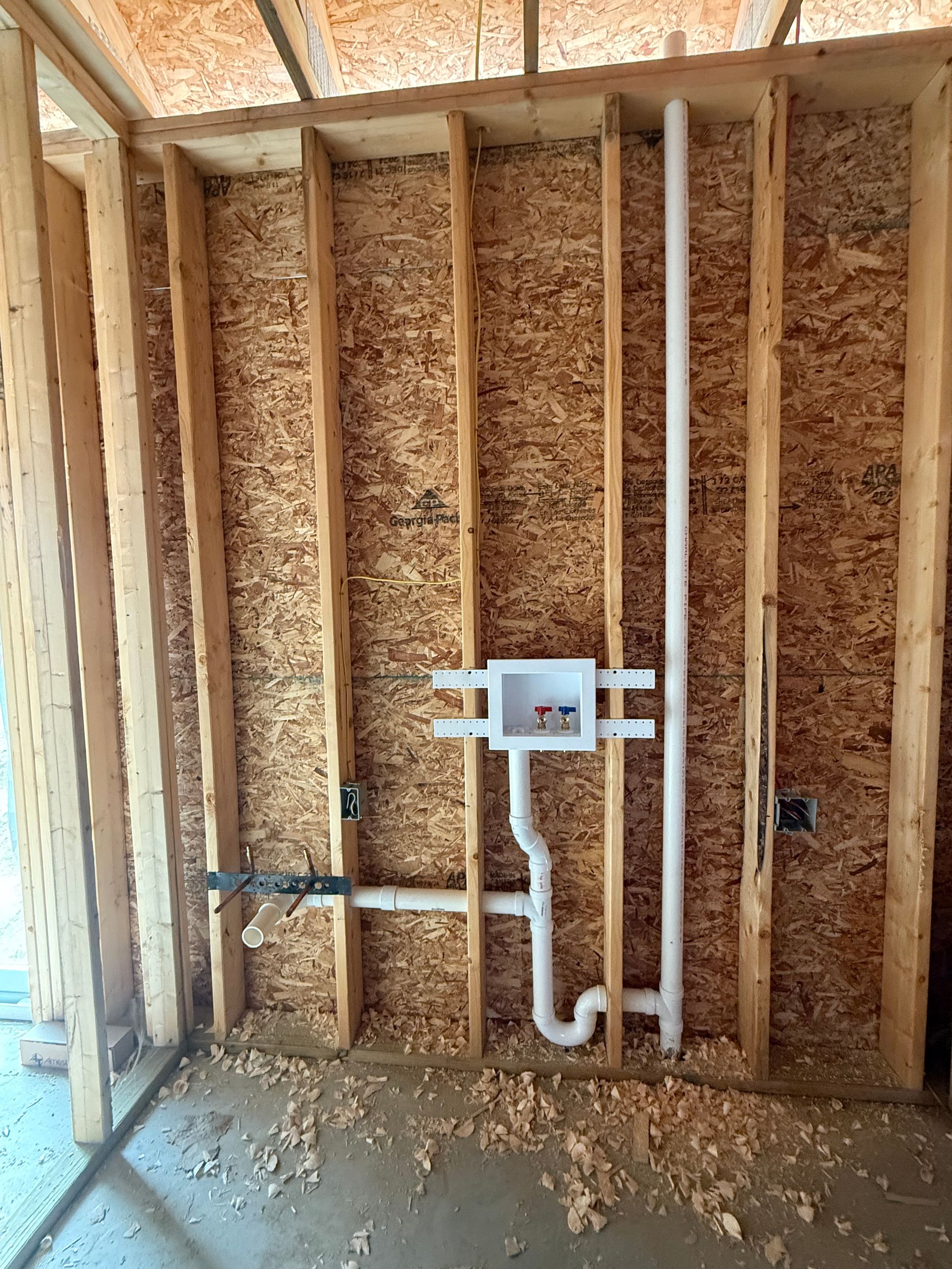 Unfinished interior wall with exposed studs, plumbing pipes, and a utility box.