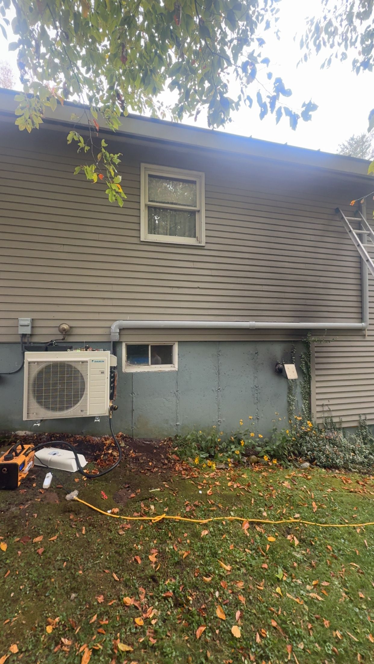 House exterior with gray siding, small window, air conditioner, and fallen leaves under overhanging tree branches