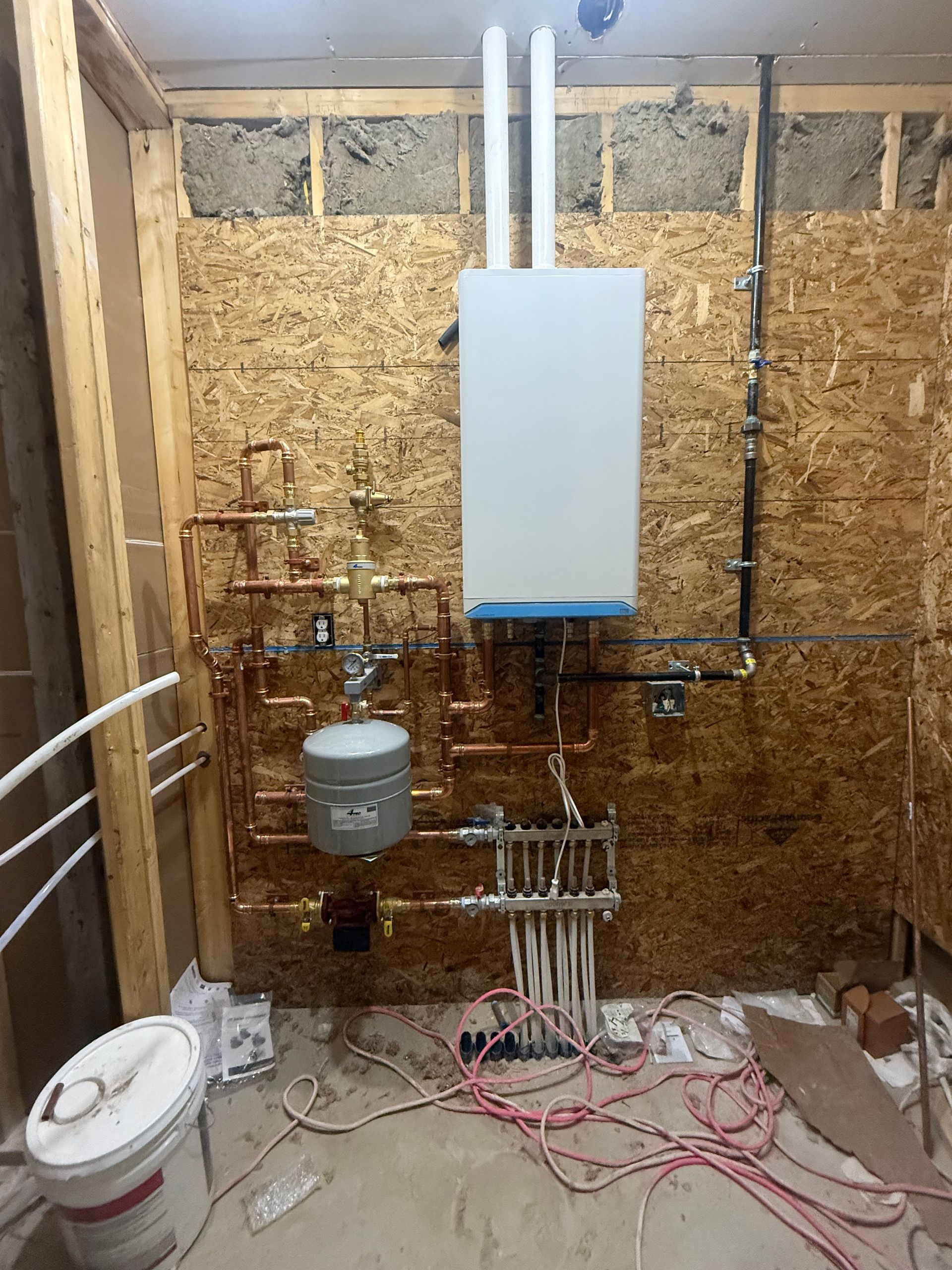Utility room with exposed plumbing, water heater, pipes, and tangled cables against unfinished wood walls.