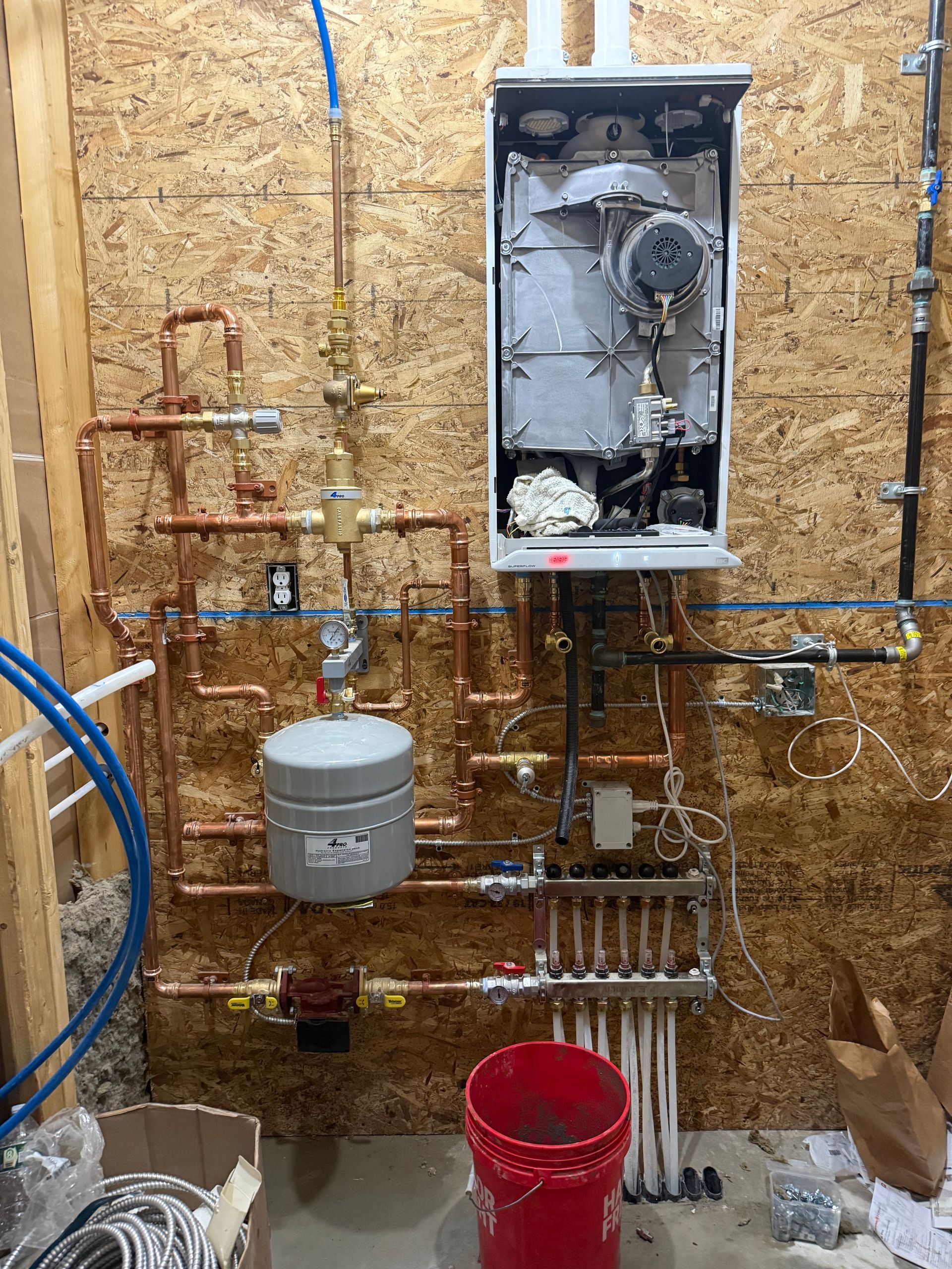 Open utility wall with exposed plumbing, pipes, valves, a gray tank, and a red bucket beneath a boiler panel