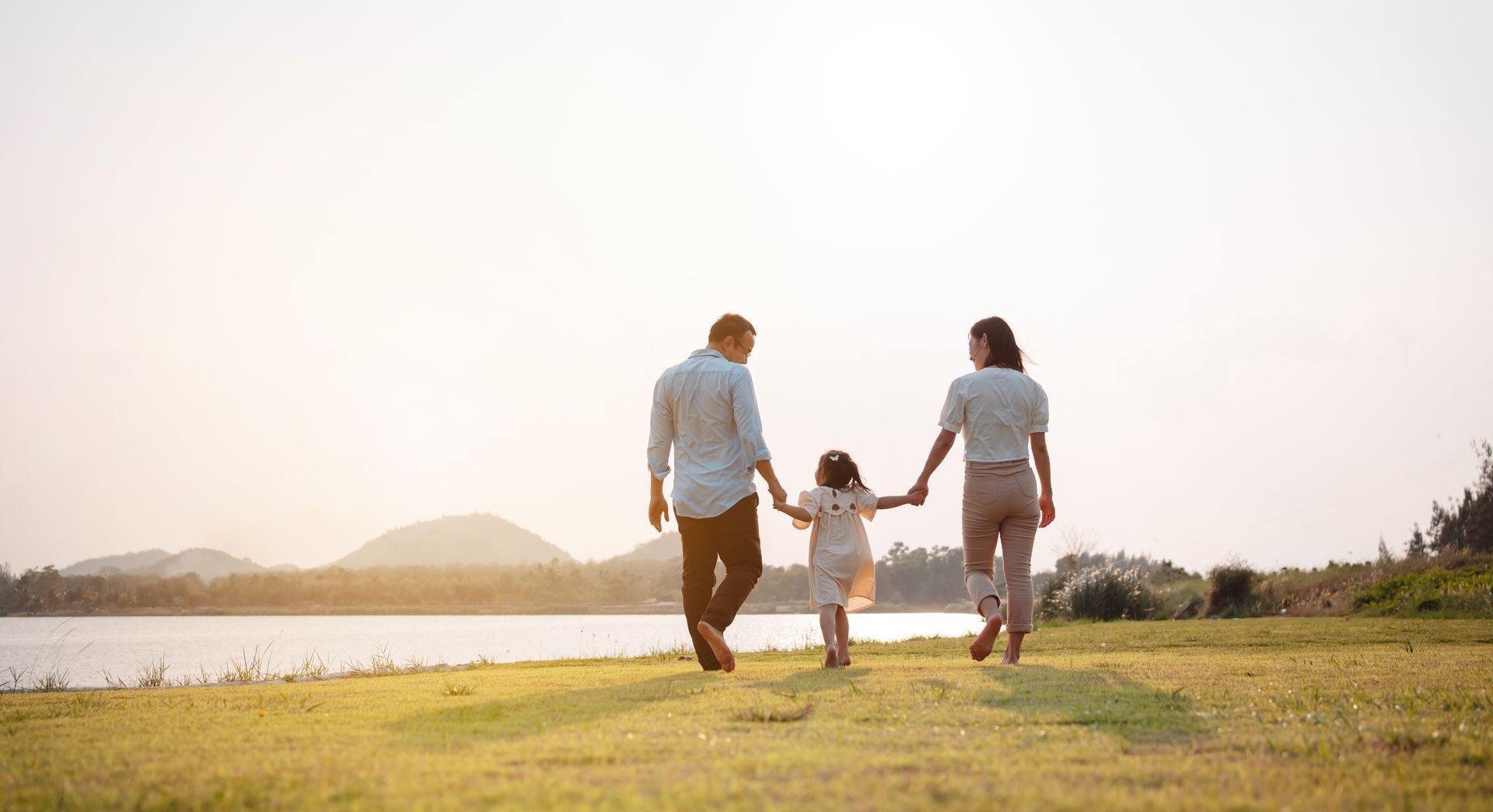 Family of three walking along a grassy bank, holding hands. The sun is setting over a lake.