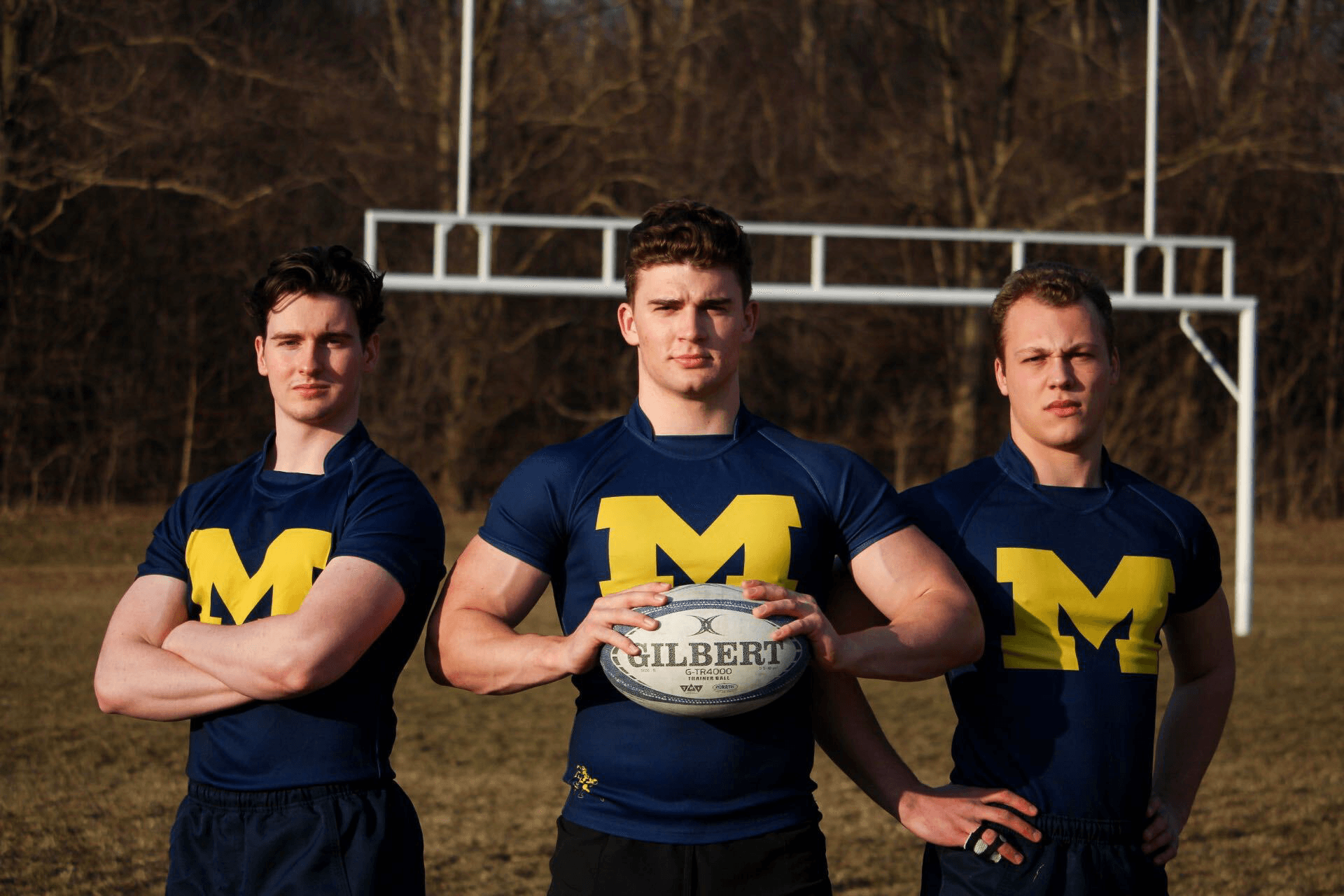 Three men wearing blue shirts with the letter m on them holding a rugby ball