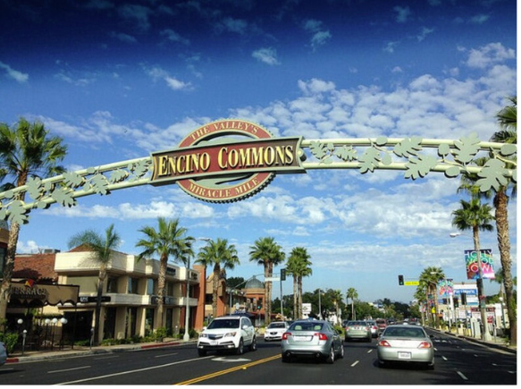 Encino Commons arched sign over street with cars, palm trees, buildings, and blue sky.