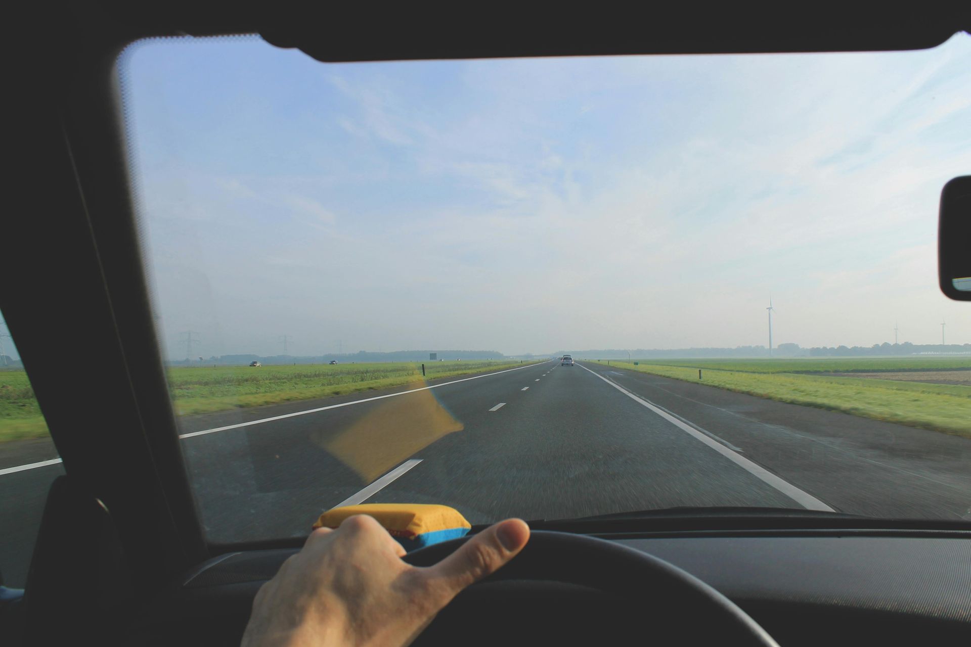 Driver’s view of a straight highway through flat fields under a clear sky, hand on the steering wheel.