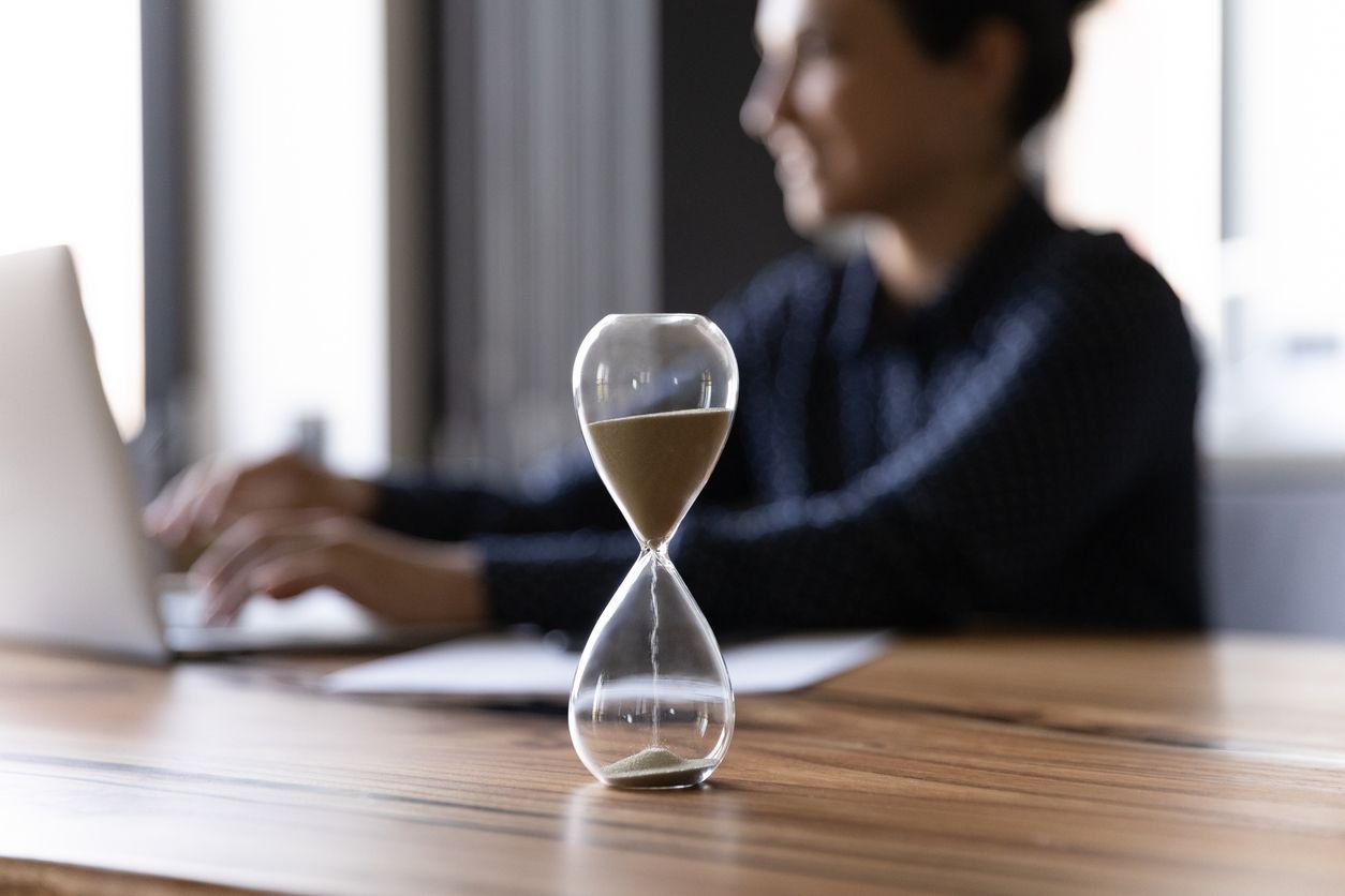 A man is typing on a laptop next to an hourglass on a table.