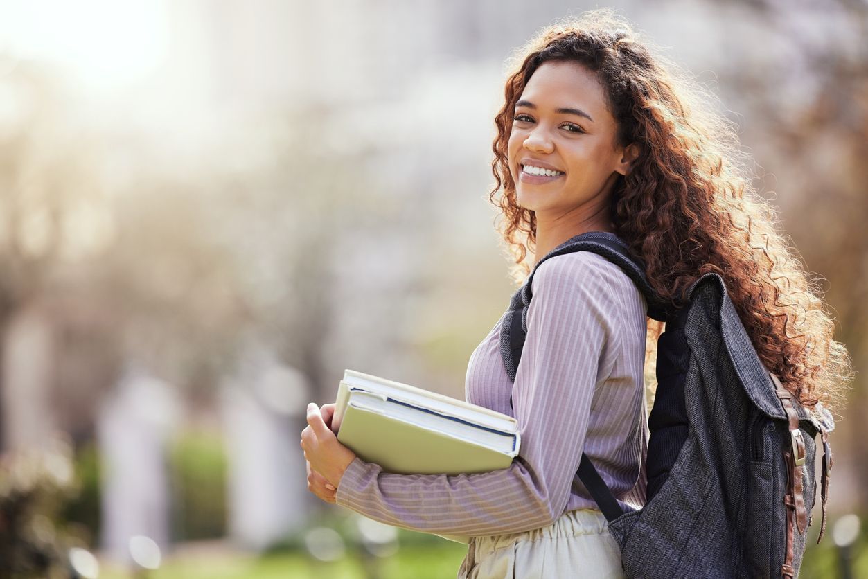 A woman with curly hair is holding a book and a backpack.