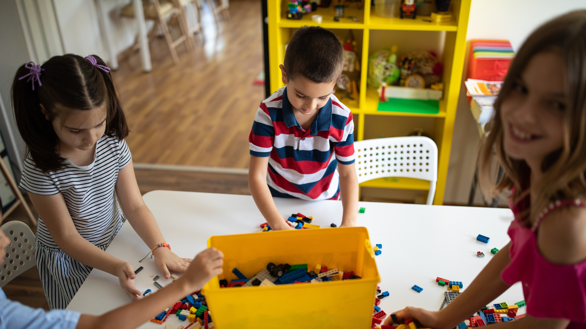 Children playing with colorful building blocks at a table. A yellow bin holds the blocks.