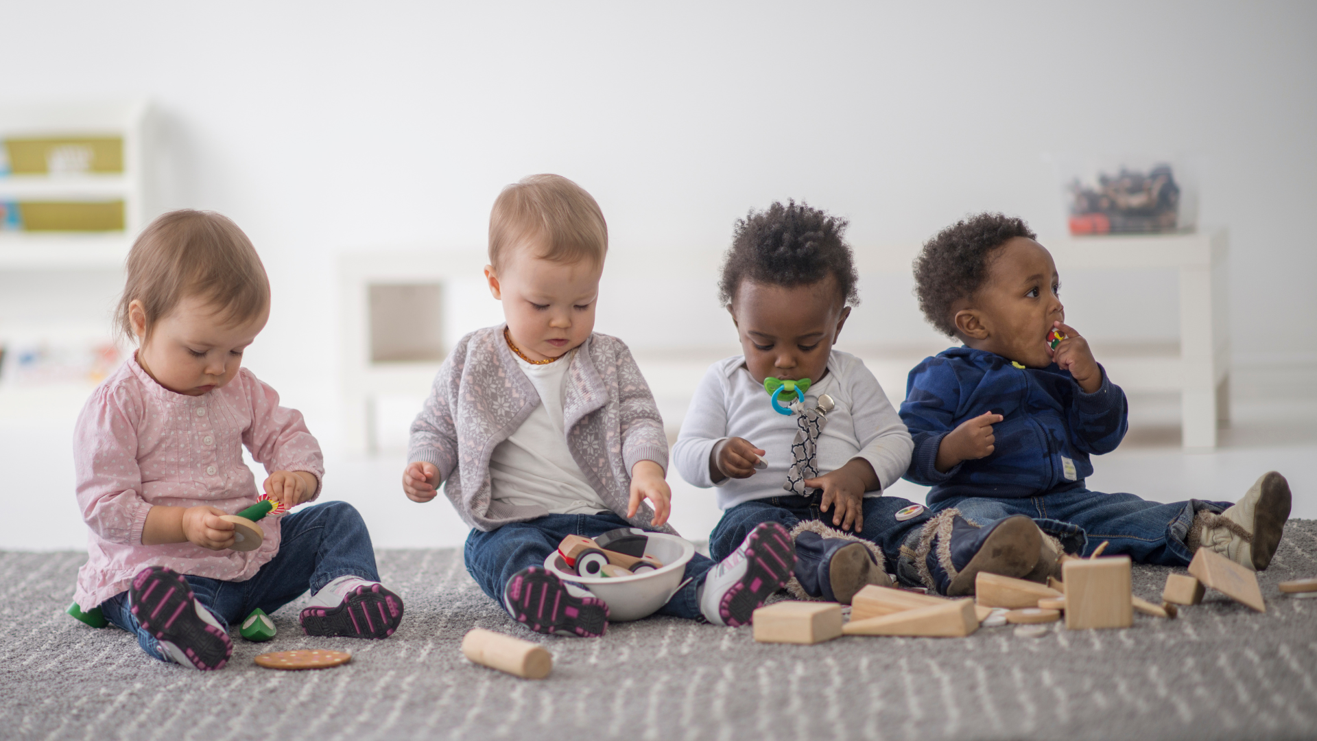 Four babies of different ethnicities playing on a rug indoors with toys.