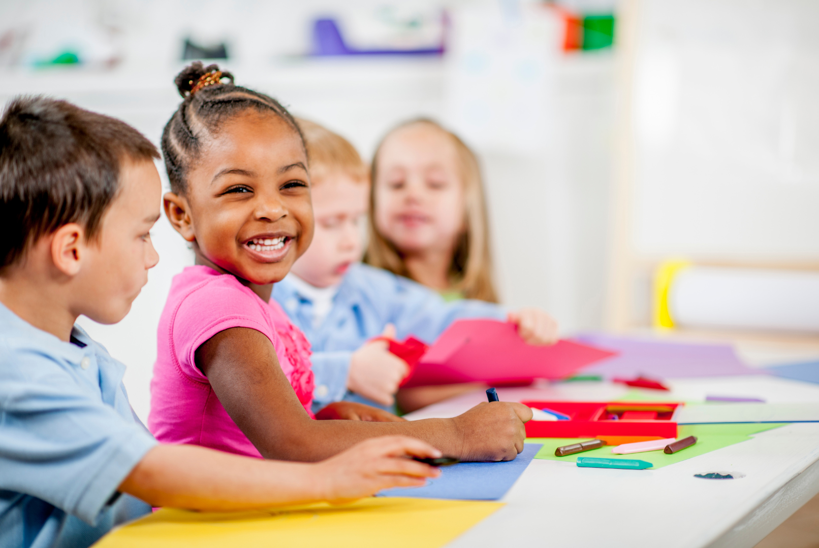 Woman and child drawing with crayons at a table, another child and adult in the background in a playroom.