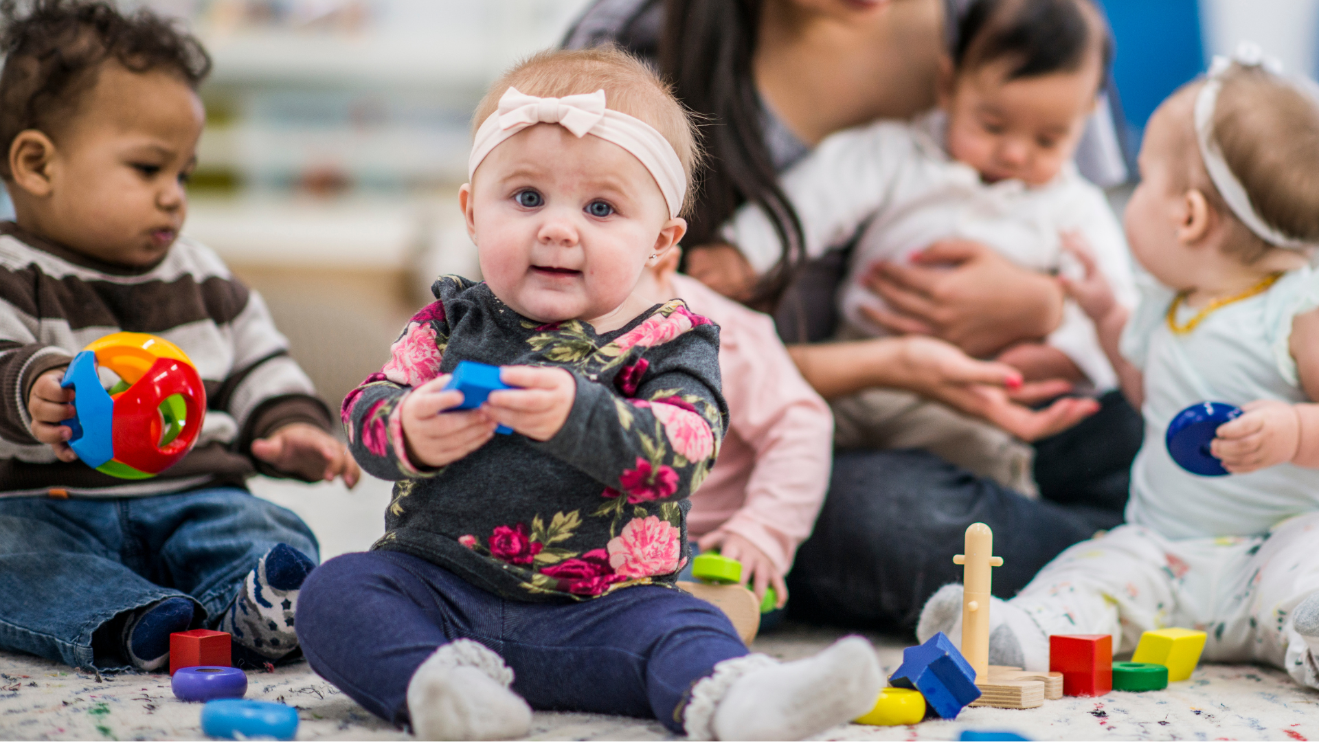 Woman assists a baby on a mat with toys in a gym. The woman smiles.