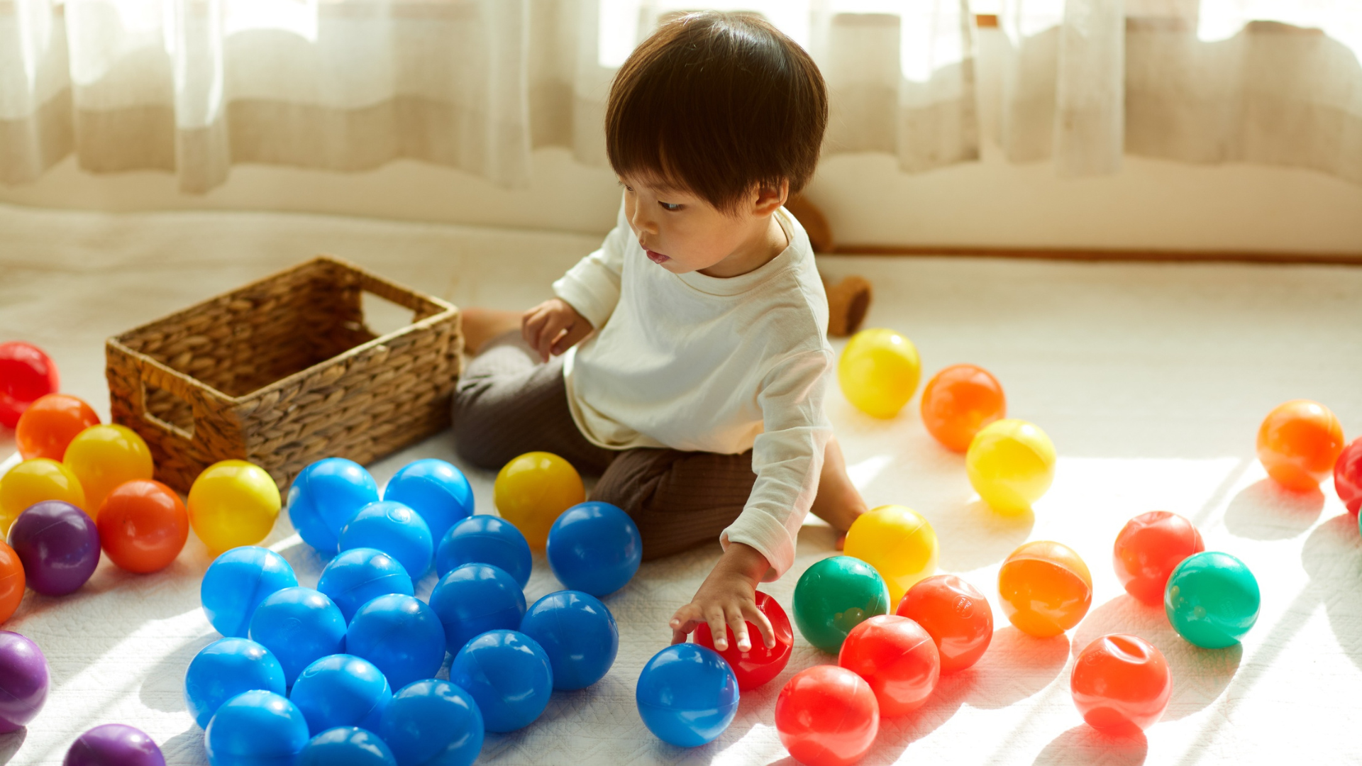 A young child sits among colorful balls, sorting them on the floor in a sunny room.