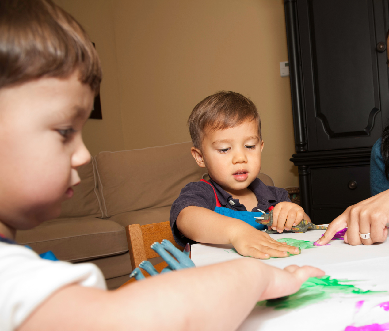Two young children playing with green play dough at a table; an adult's hand points.