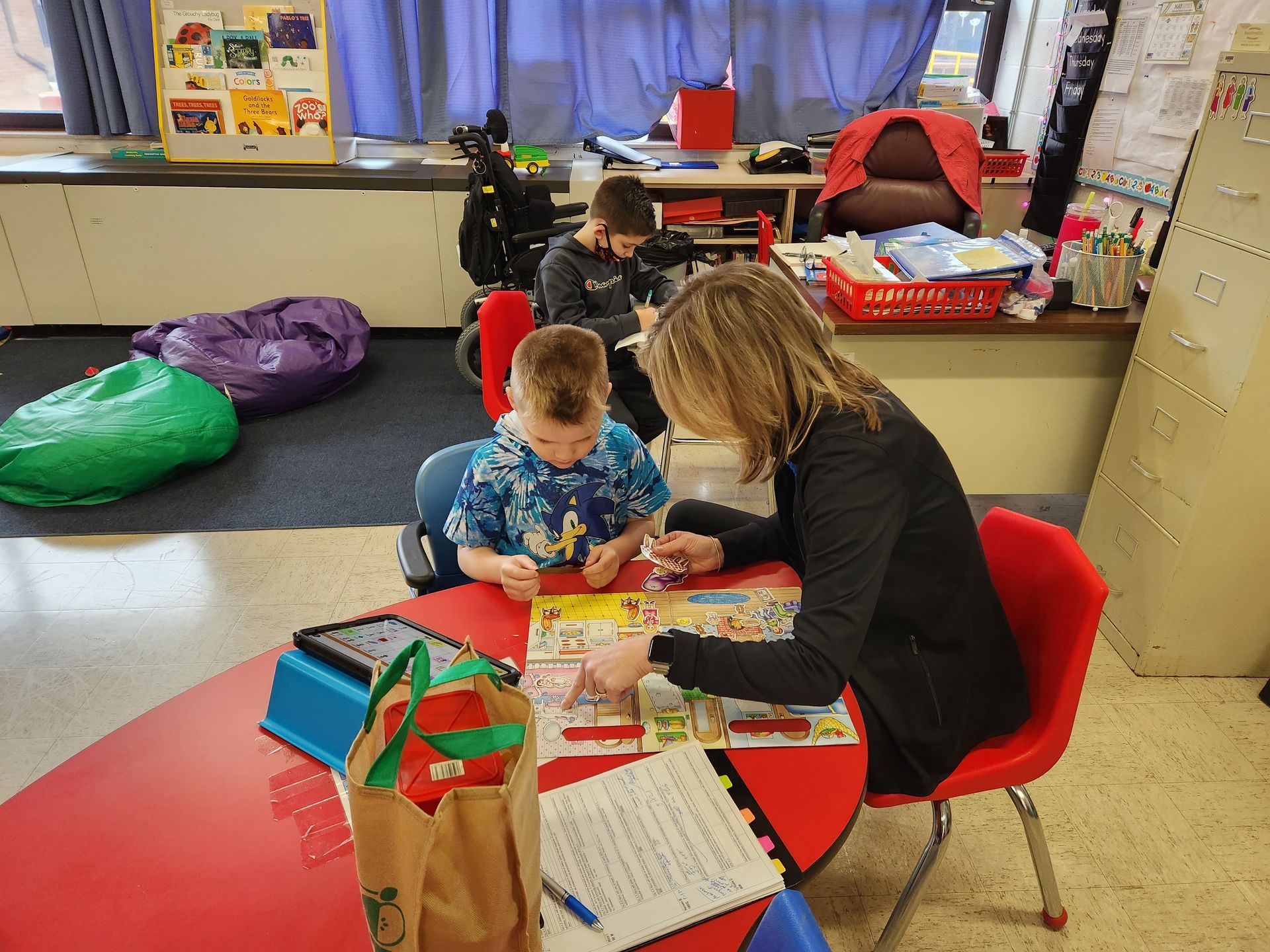 A teacher assists two students at a red table in a classroom.