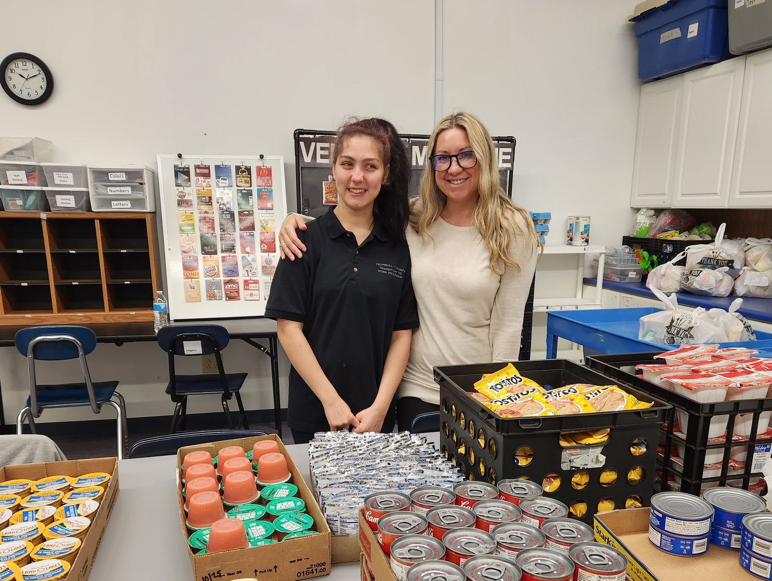 Two women stand behind a table with food items. One has an arm around the other. Shelf in background.