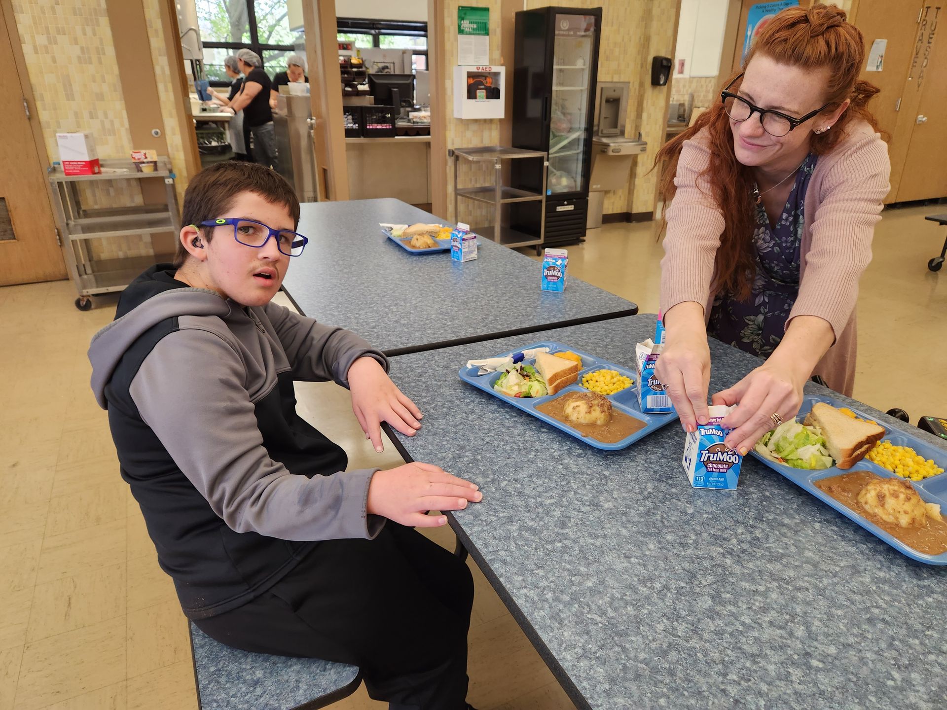 A person assists a seated person at a lunch table with a tray. Indoor setting with prepared meals.