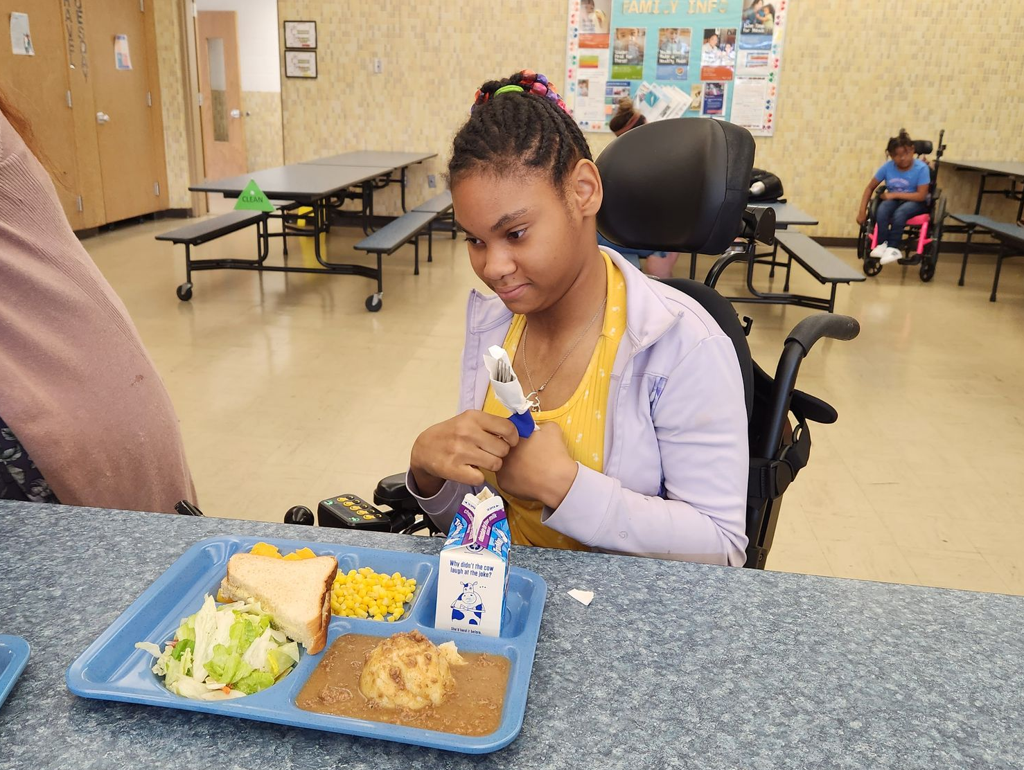 Girl in a wheelchair at a cafeteria table eating a meal. Tray with food and drink.