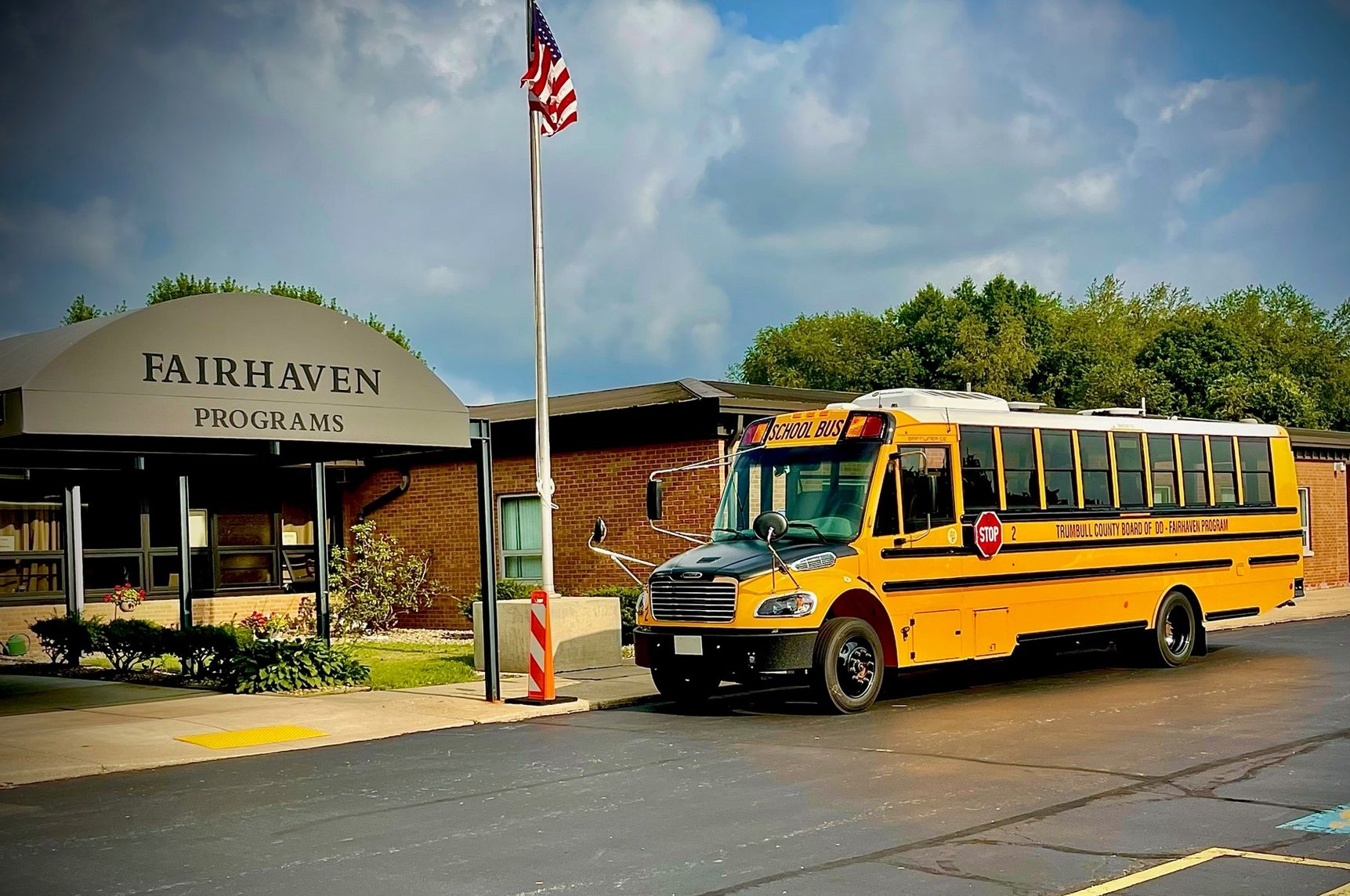 Yellow school bus parked in front of Fairhaven Programs building with an American flag waving in the sky.