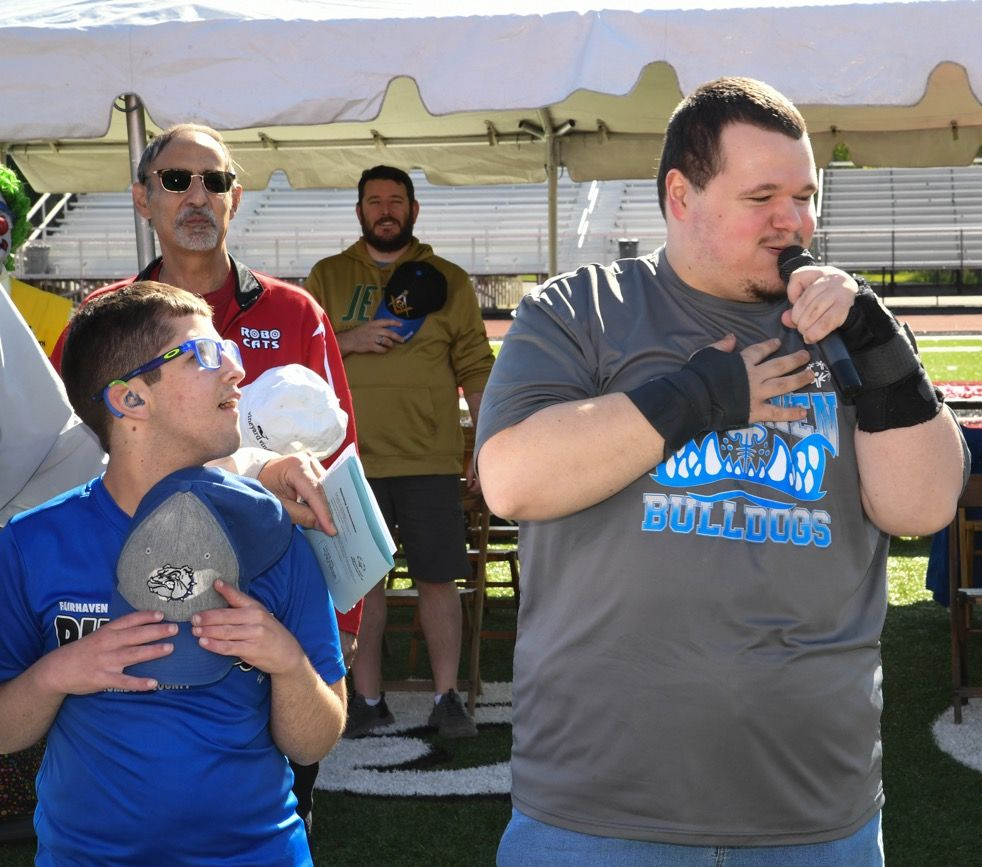 Two men, one speaking into a mic with hand on chest, other holding a cap. Two men behind them. Outdoors, sunny.