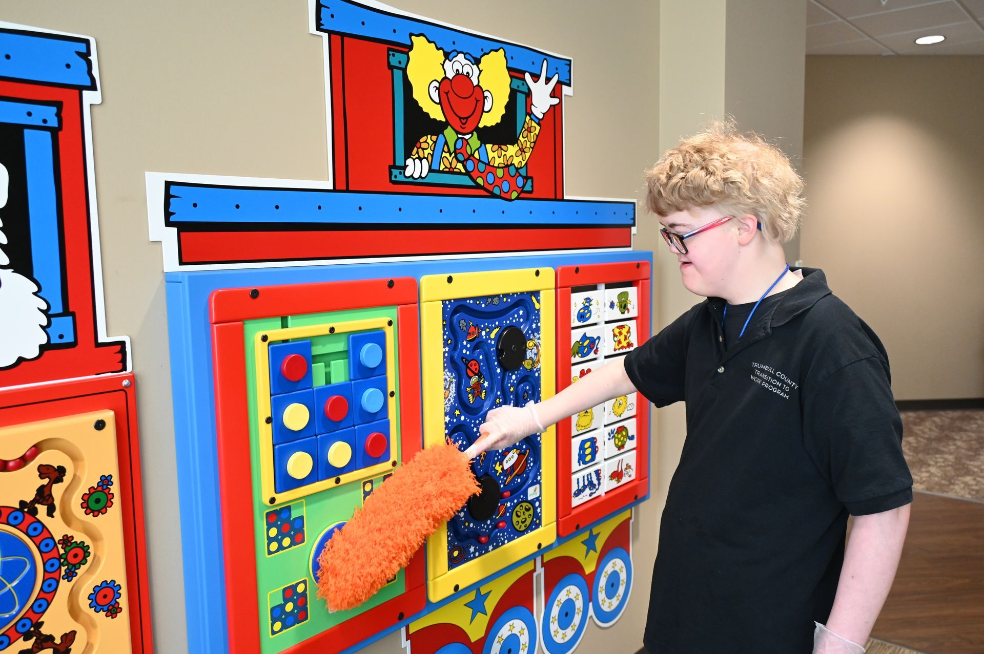 A person with Down syndrome uses an orange cloth to clean a colorful wall-mounted play panel with interactive elements.