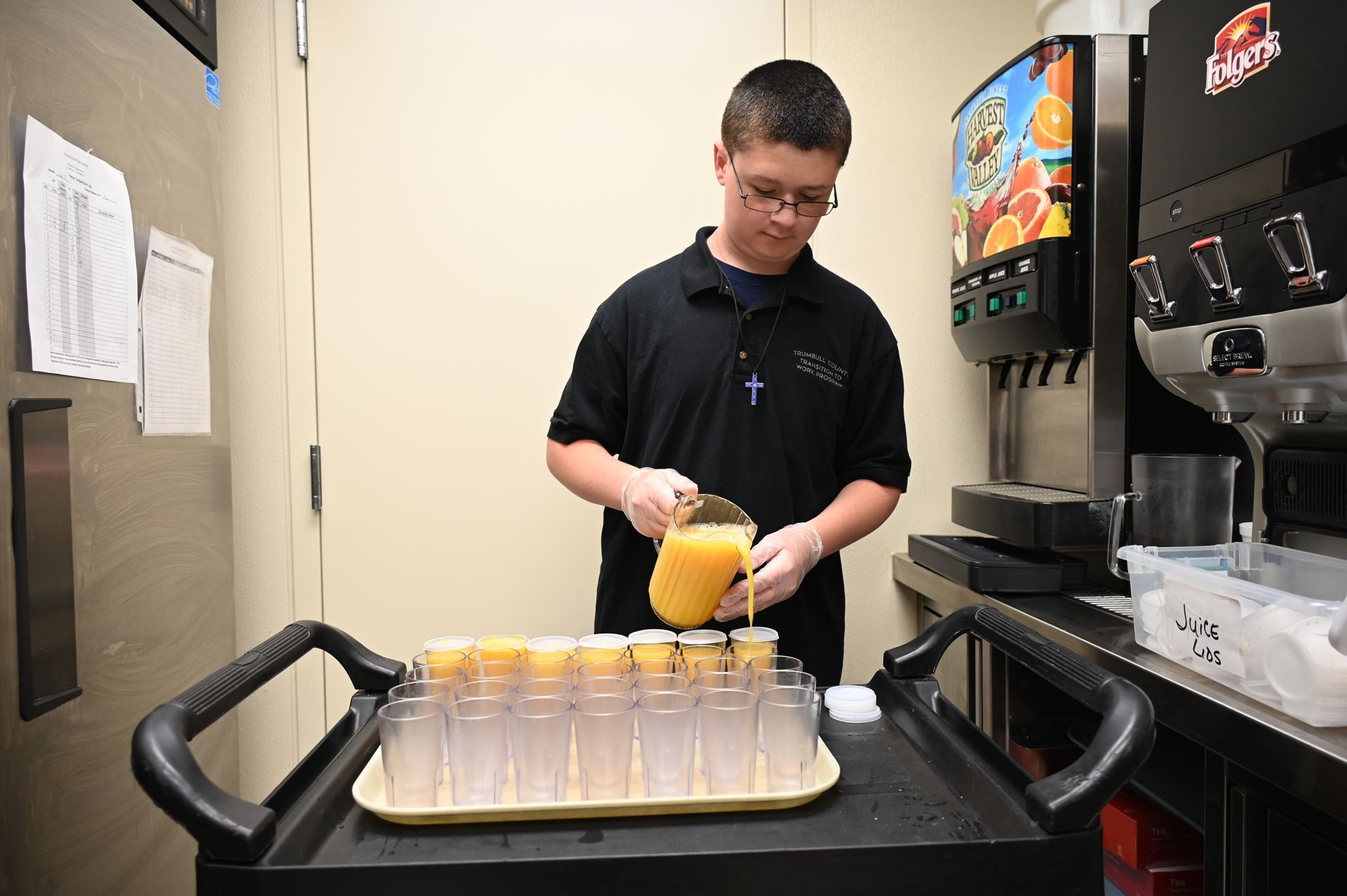 Person in black shirt pours orange juice into glasses on a tray. Juice dispenser is in background.
