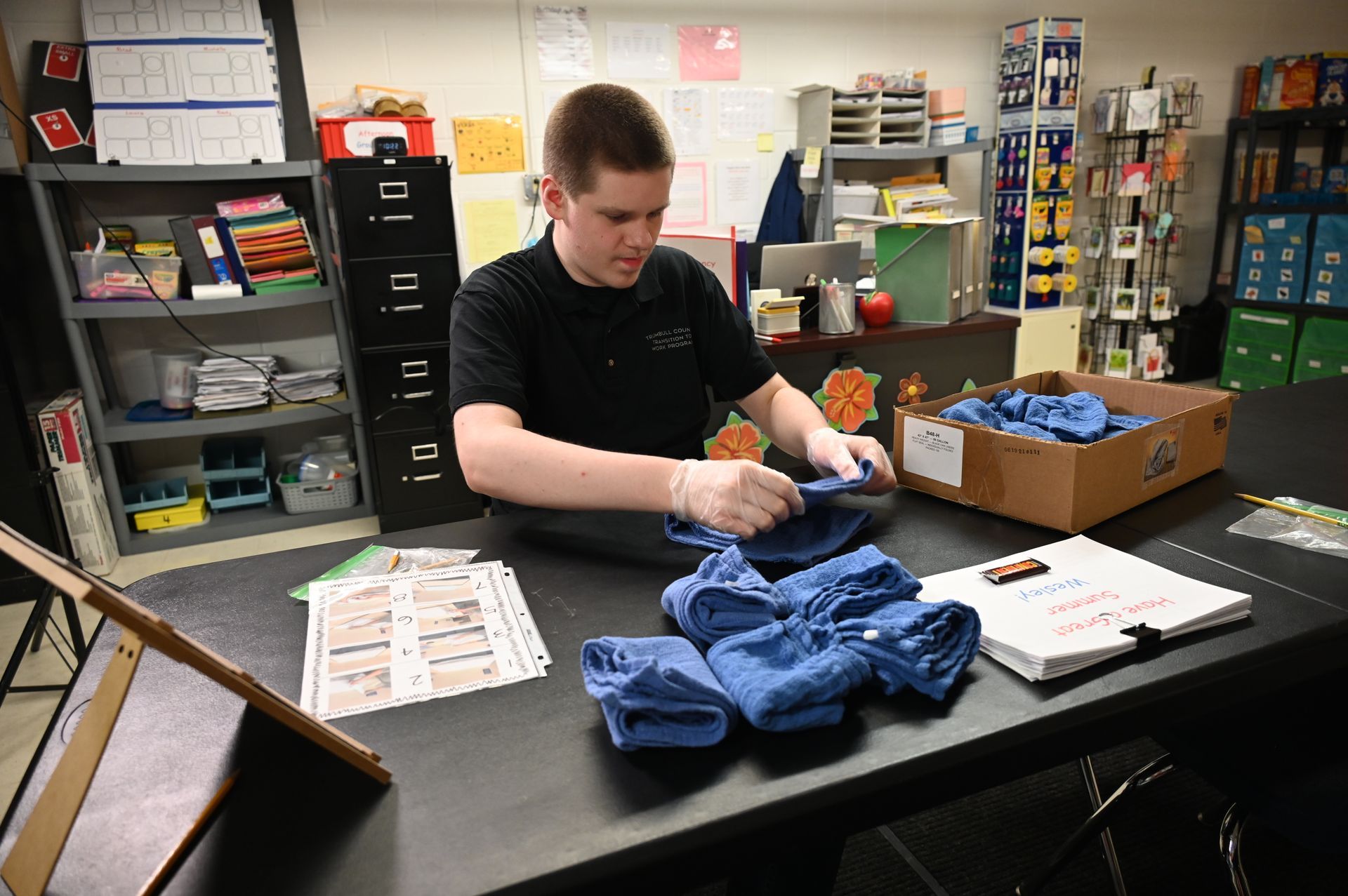 Man folding blue cloths at a table with a cardboard box, in a room with shelves and supplies.