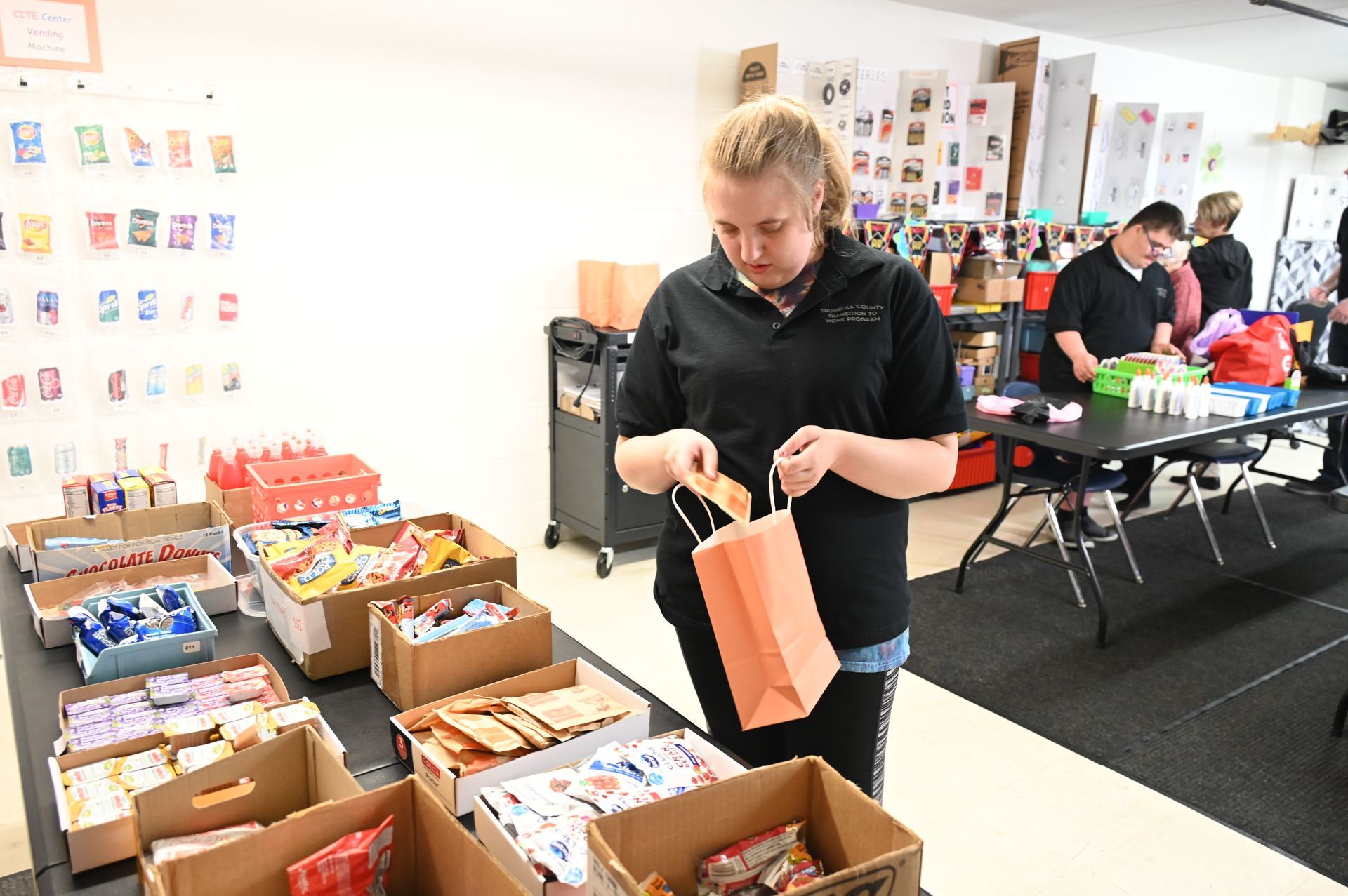 Woman filling a paper bag from boxes of snacks on a table. Others pack bags in the background. White wall.