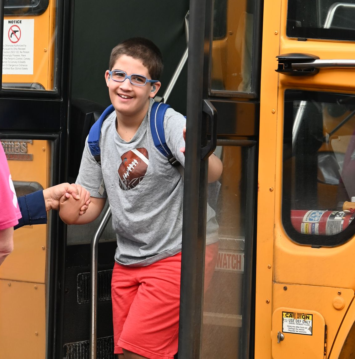 Boy with a backpack smiles while exiting a school bus, holding a hand for assistance.