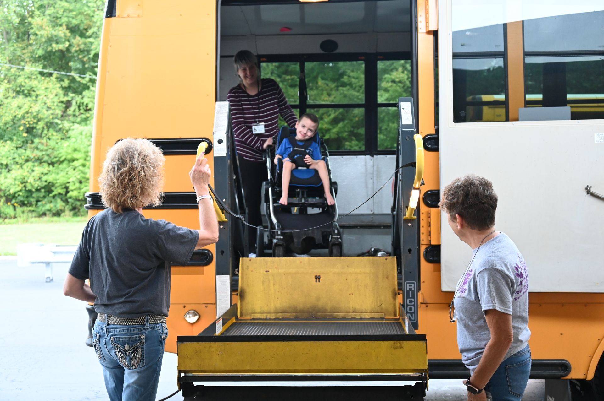 School bus with lift. A child in a wheelchair being assisted onto the bus by an adult. Two other adults watch.