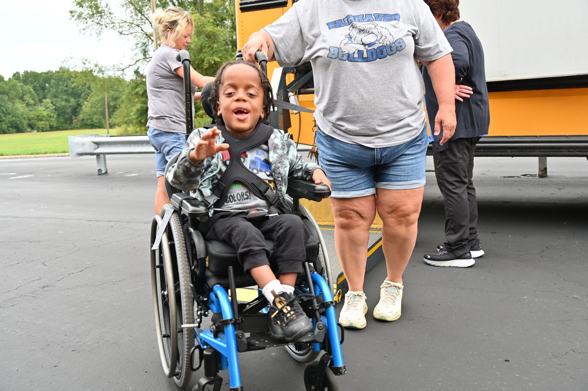 Boy in wheelchair with adults near a school bus. He smiles, points forward.