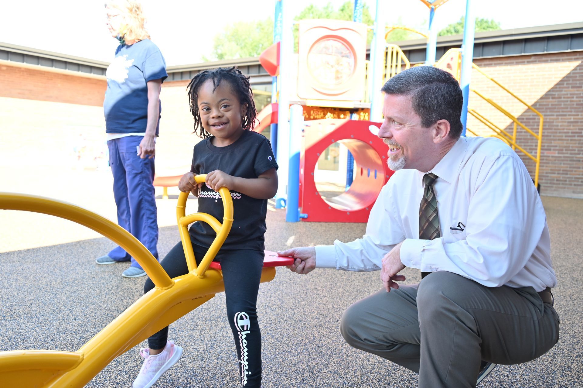 Child playing on a seesaw with an adult at a playground; another person watches.