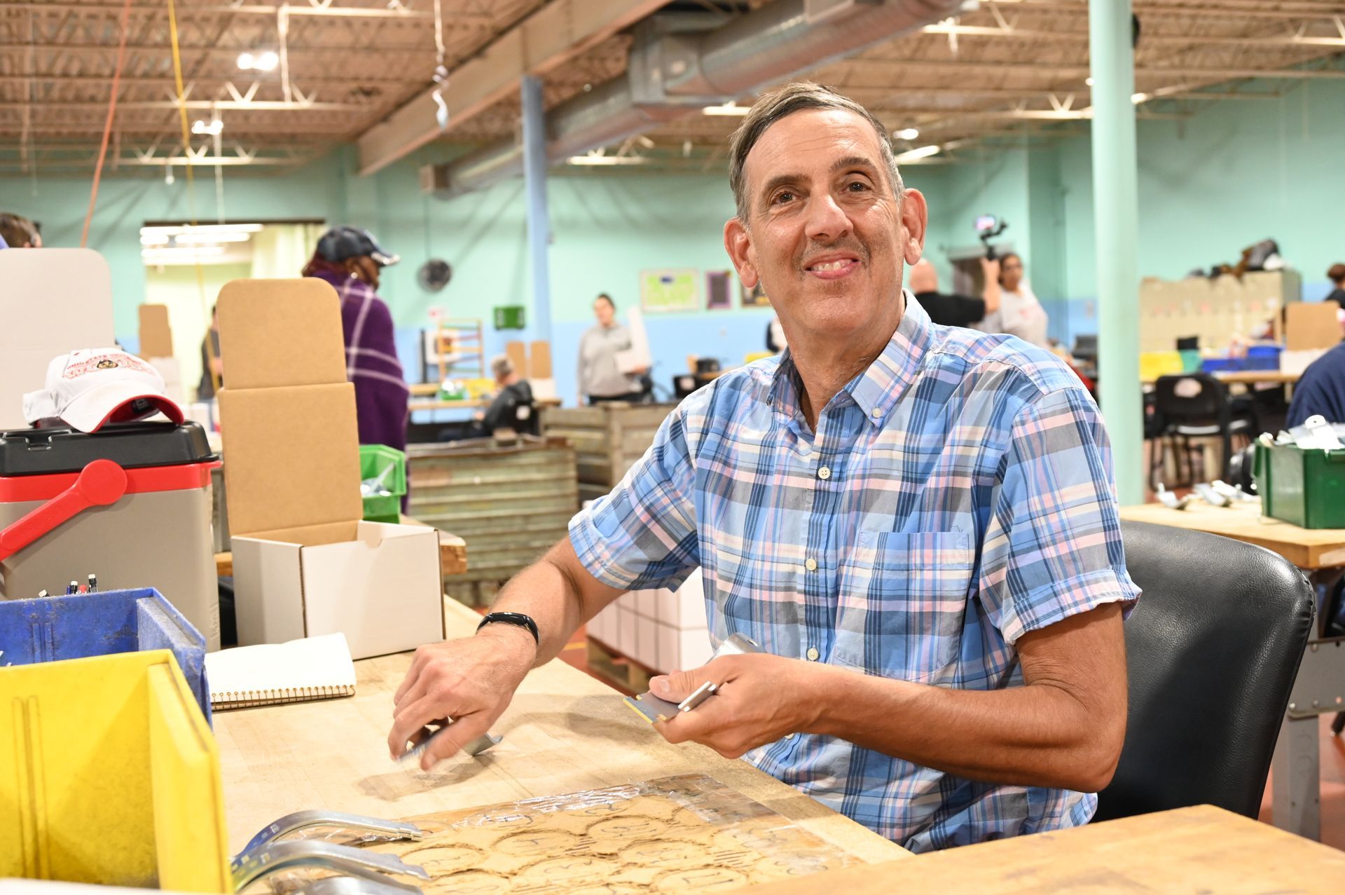 Man smiling, working at a table in a warehouse setting, wearing a plaid shirt.