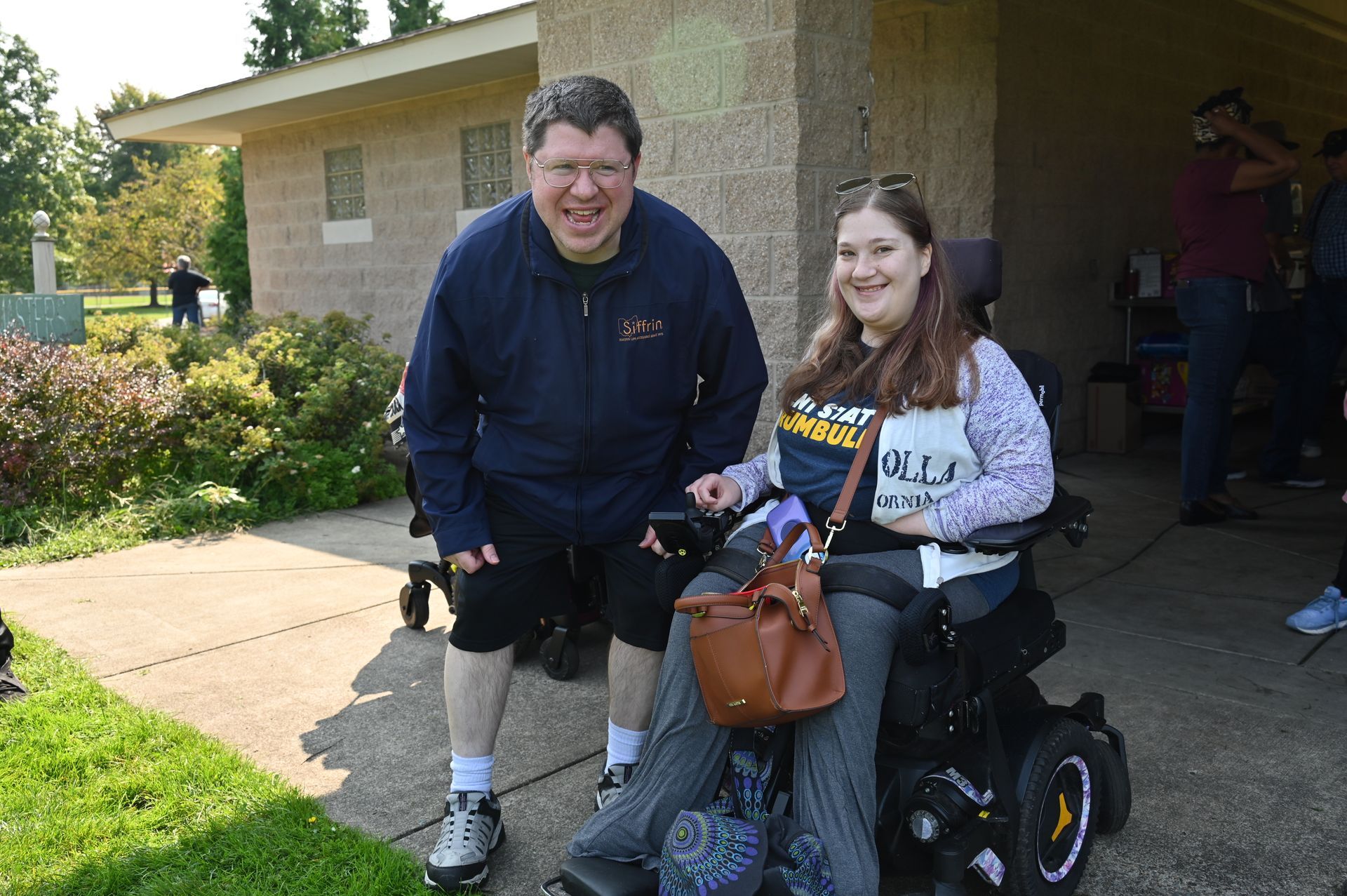 Man and woman smiling, near a building. Woman in a wheelchair, with a purse. Outside, sunny day.