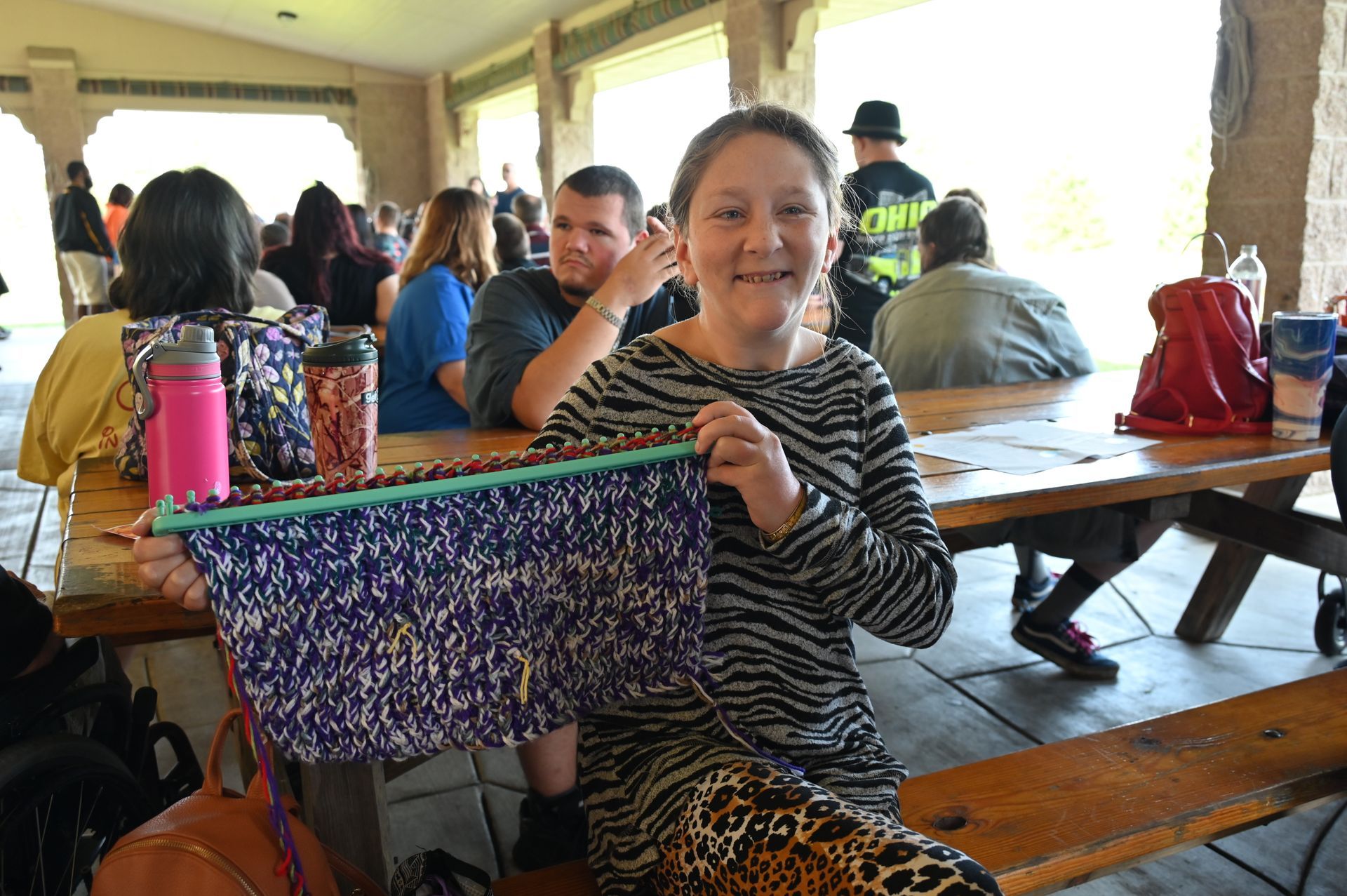 Woman holding a knitted blue, purple, and yellow project at a picnic table, smiling.