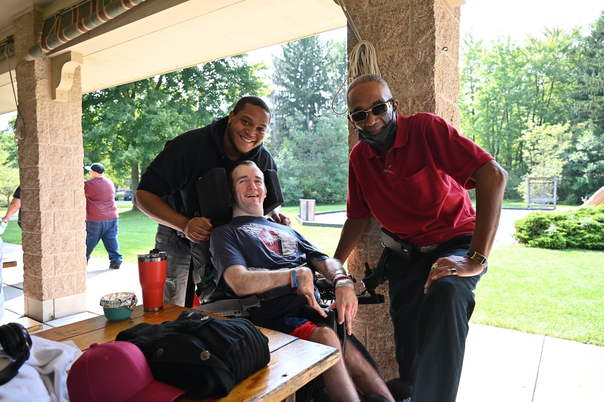 Three people pose for a photo in a park. One sits in a wheelchair, smiling. Others smile, one wears a red shirt.
