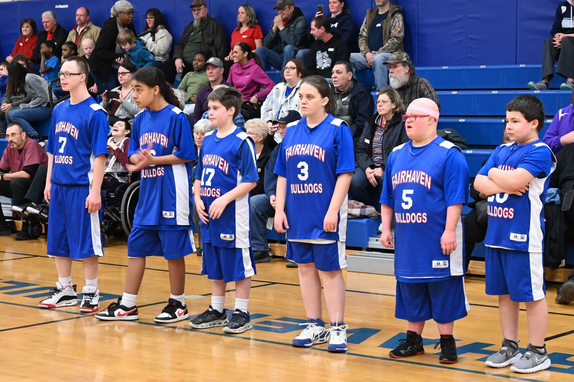 Basketball team in blue uniforms stands on a court, crowd in background.
