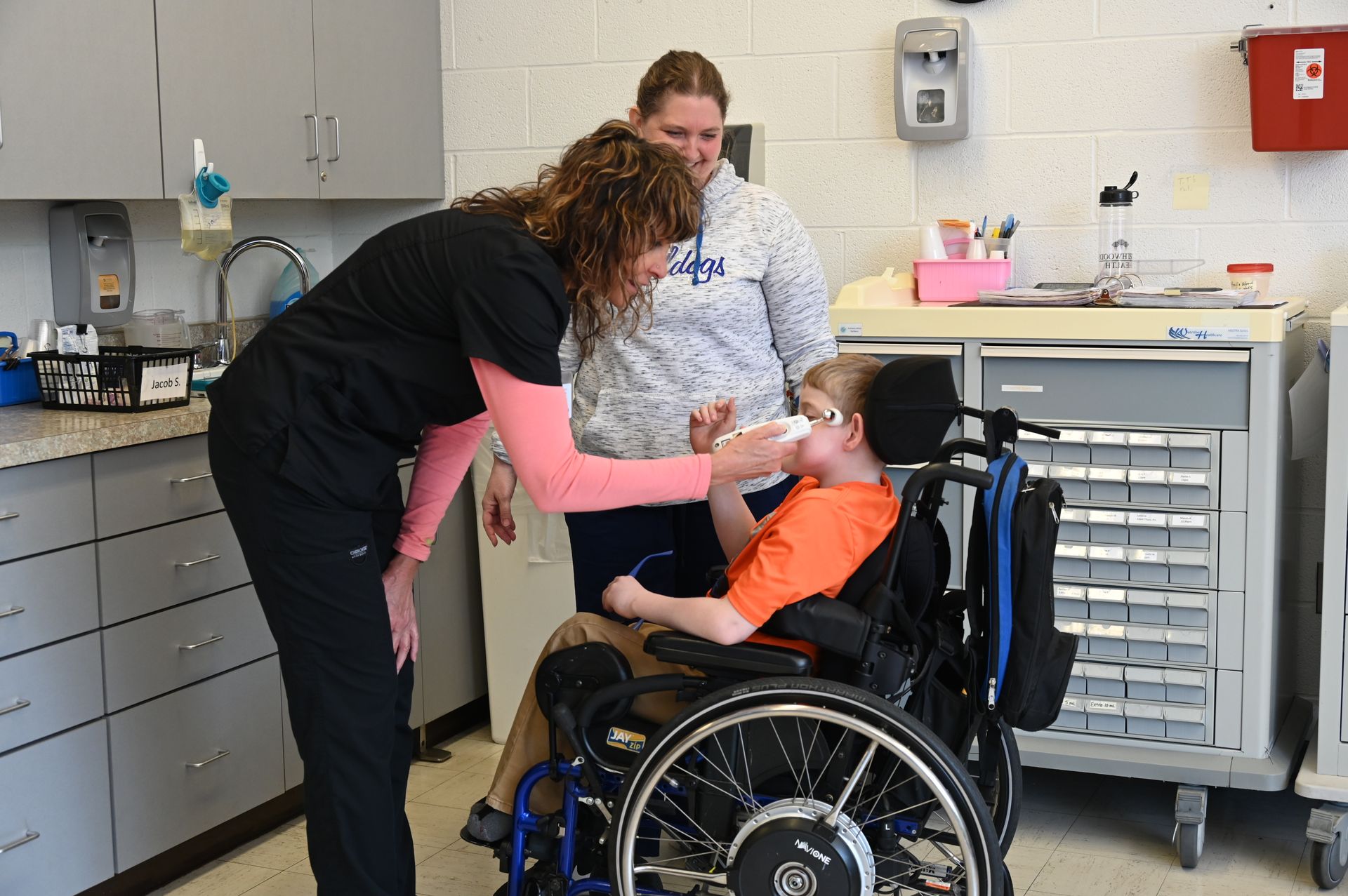 A person in scrubs assists a child in a wheelchair with something at their mouth, as an adult watches.