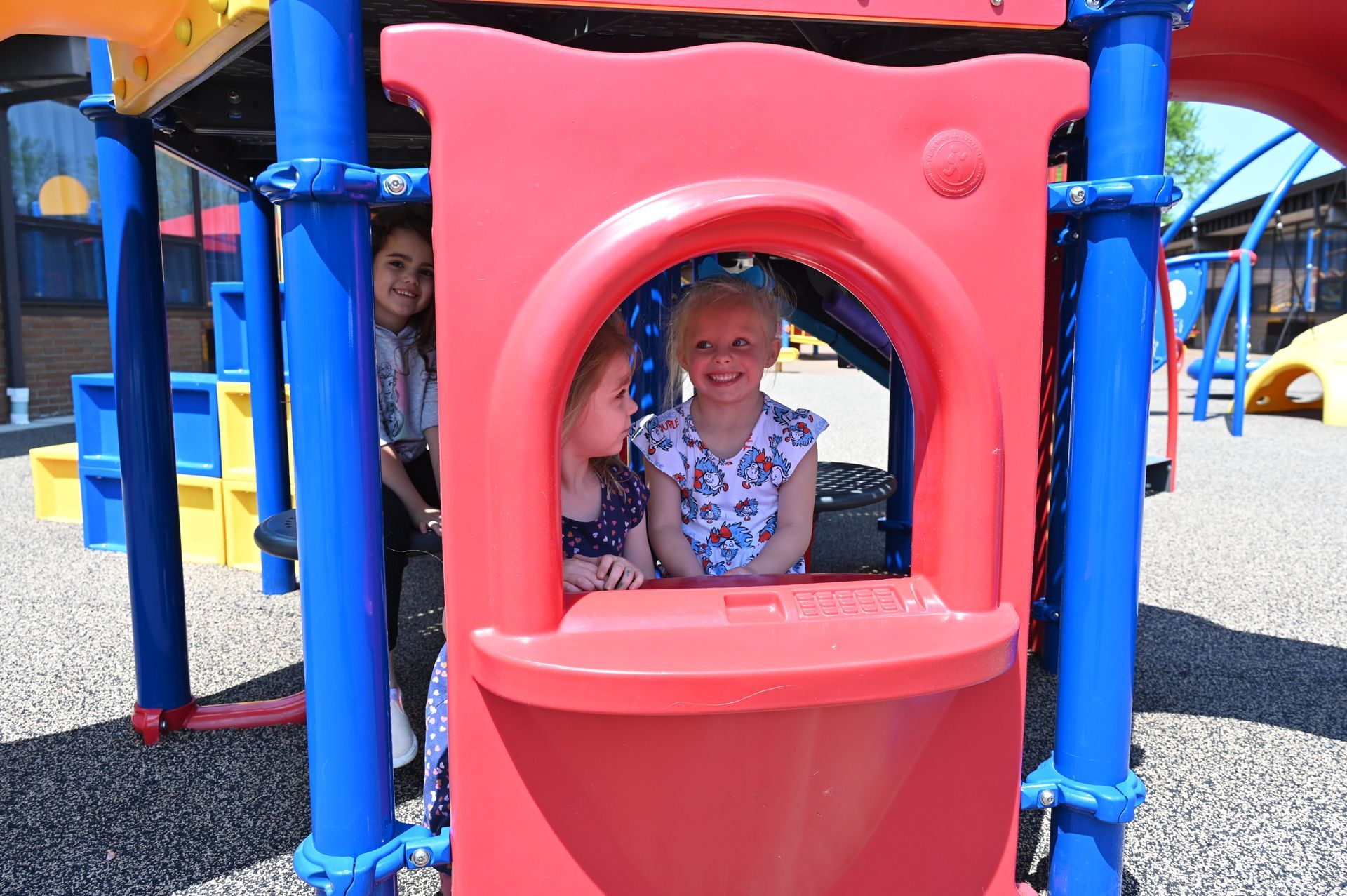 Three children playing on a red and blue playground structure, smiling.