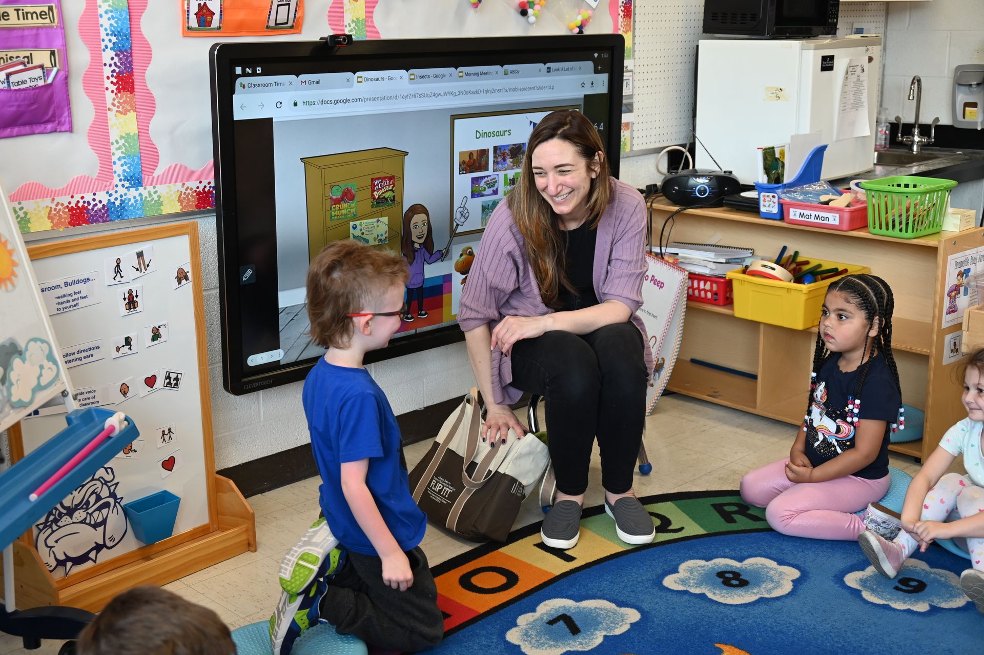 Teacher with children in a classroom; they are sitting on a rug and looking at a digital screen.