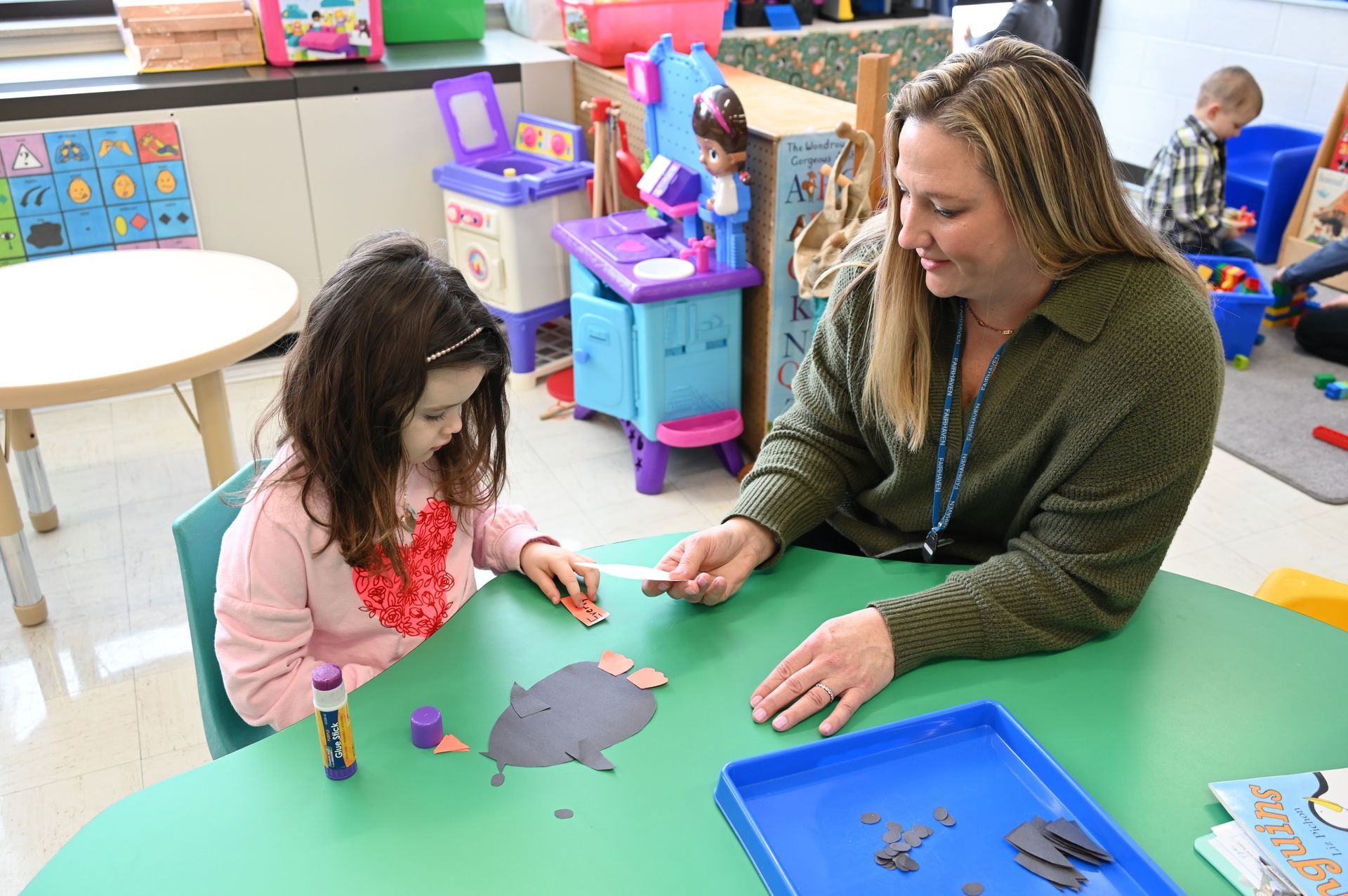 A woman and a young child crafting at a table in a brightly lit room with toys.