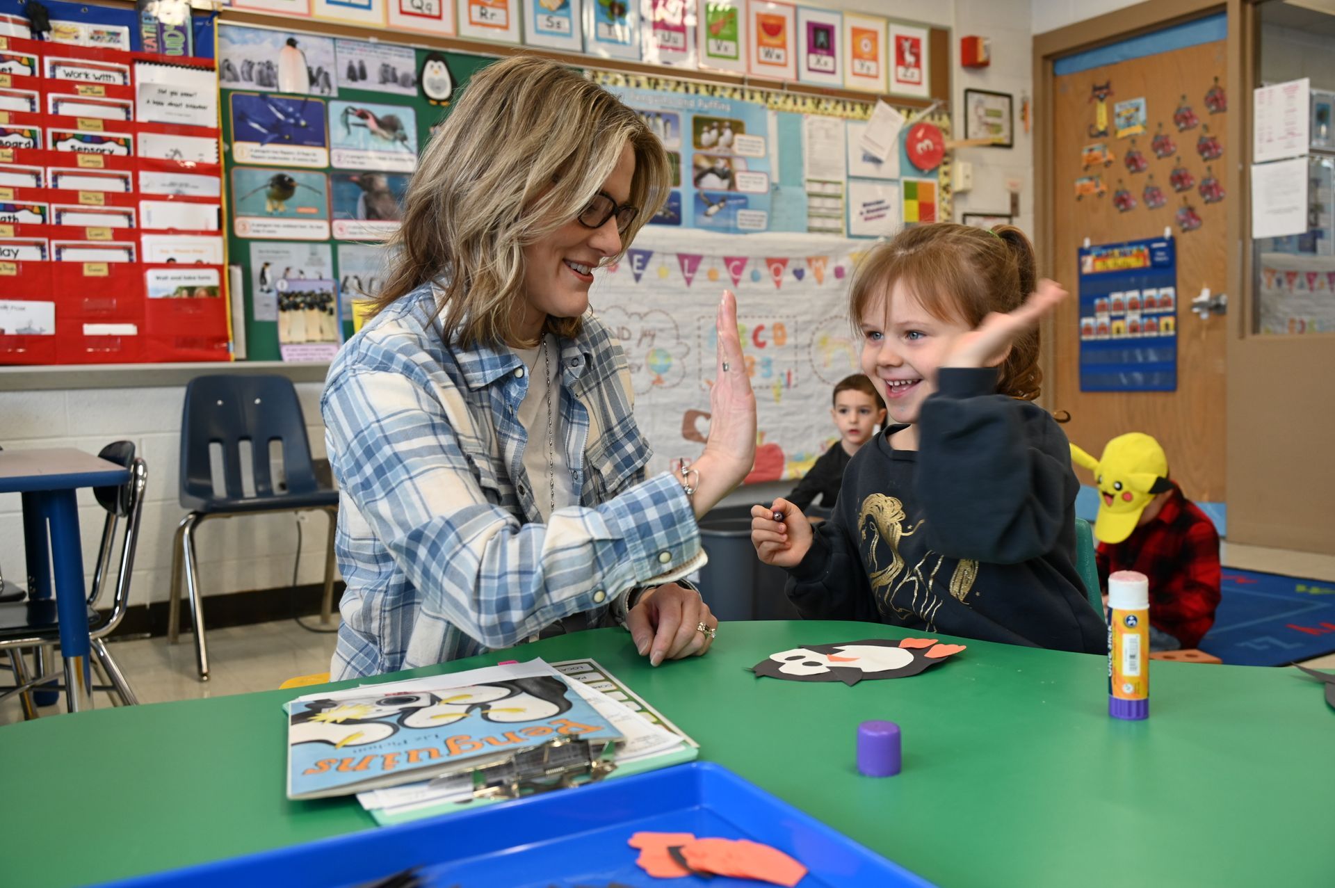 Teacher high-fives a smiling child at a green table in a colorful classroom. A book and glue stick are on the table.