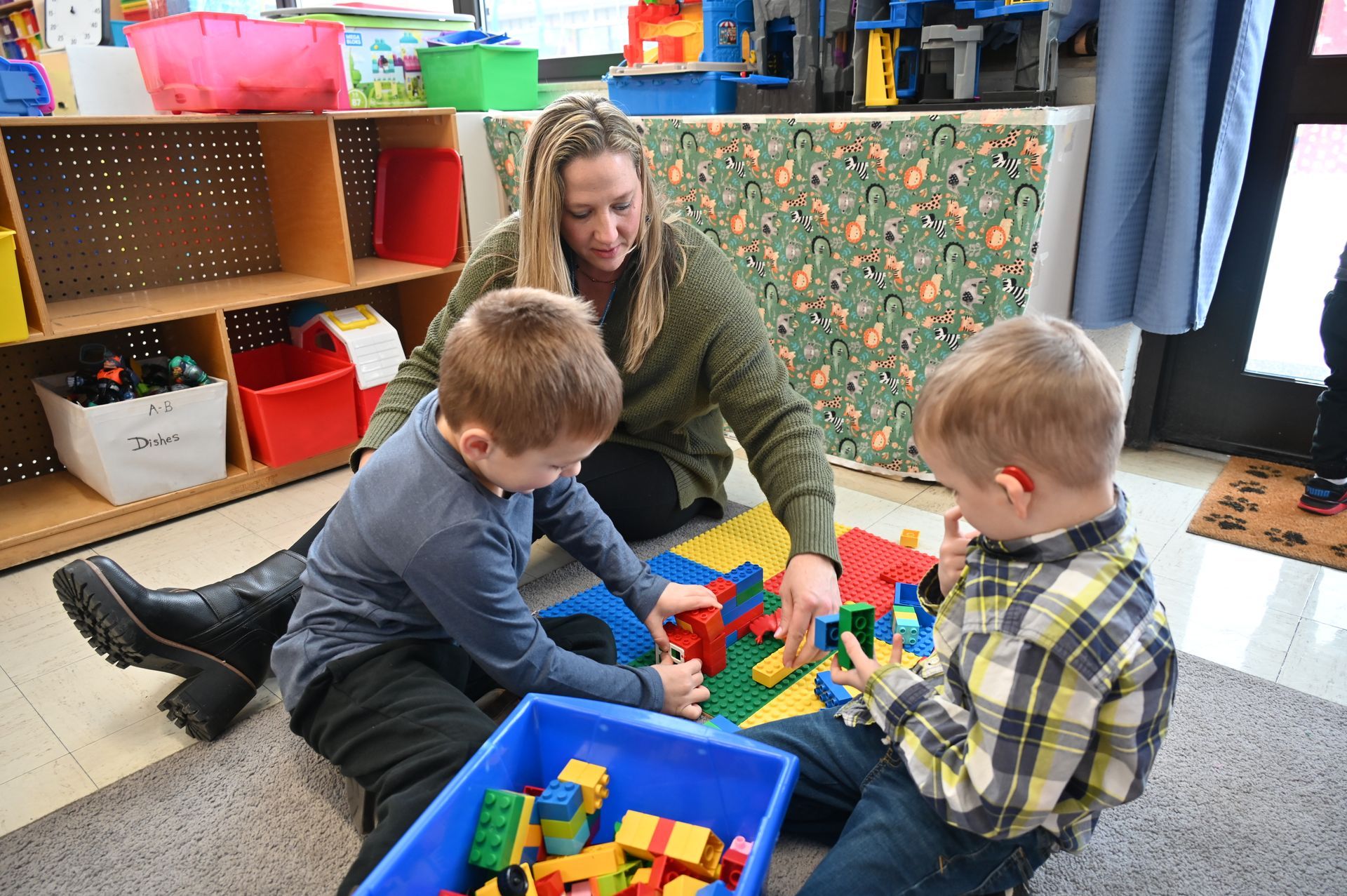 A techer sits on the floor with two children who are playing with toys. 