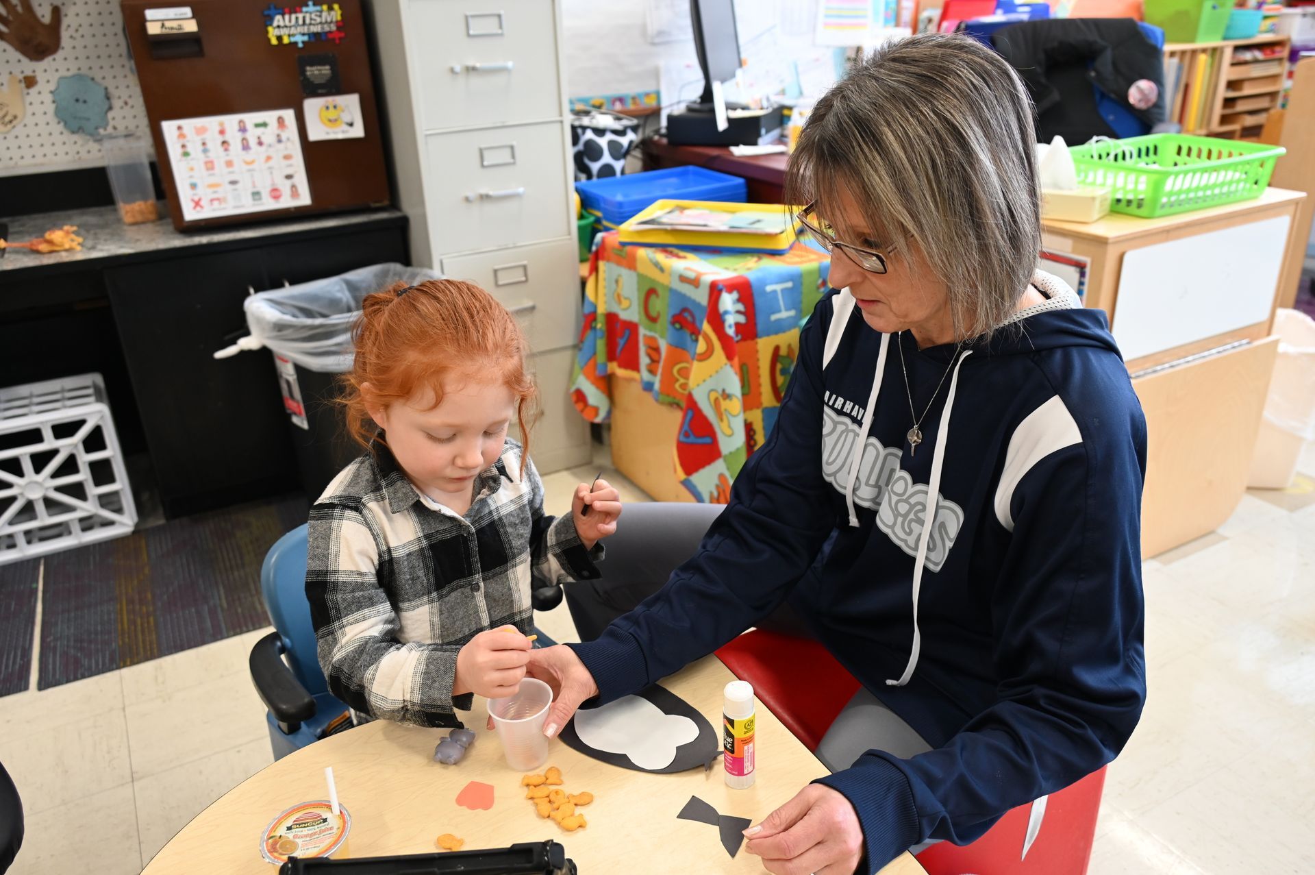 A child with red hair and an adult work on a craft project together at a table in a classroom.