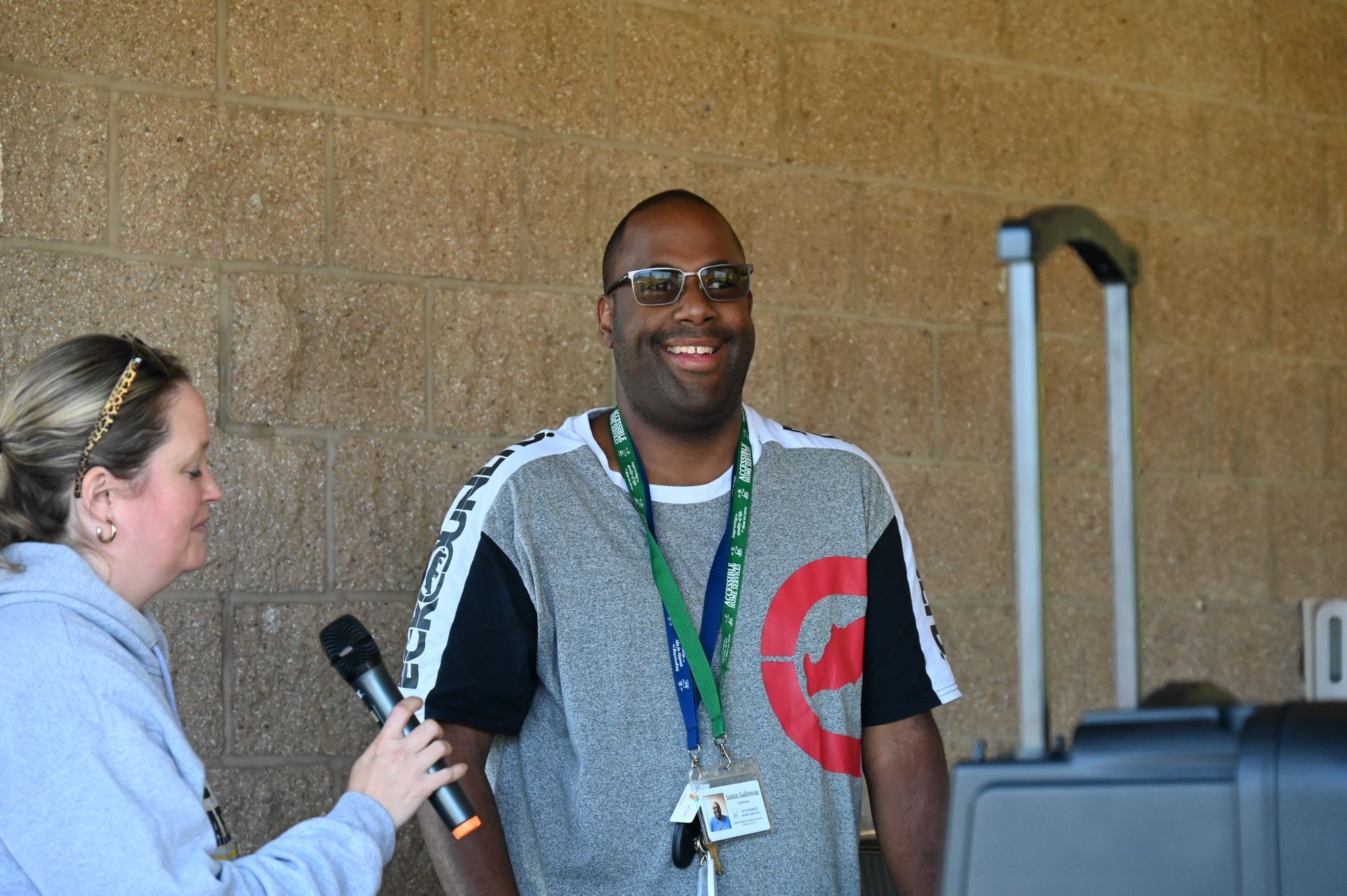A person with glasses smiles while being interviewed by a person holding a microphone. Outside a building.