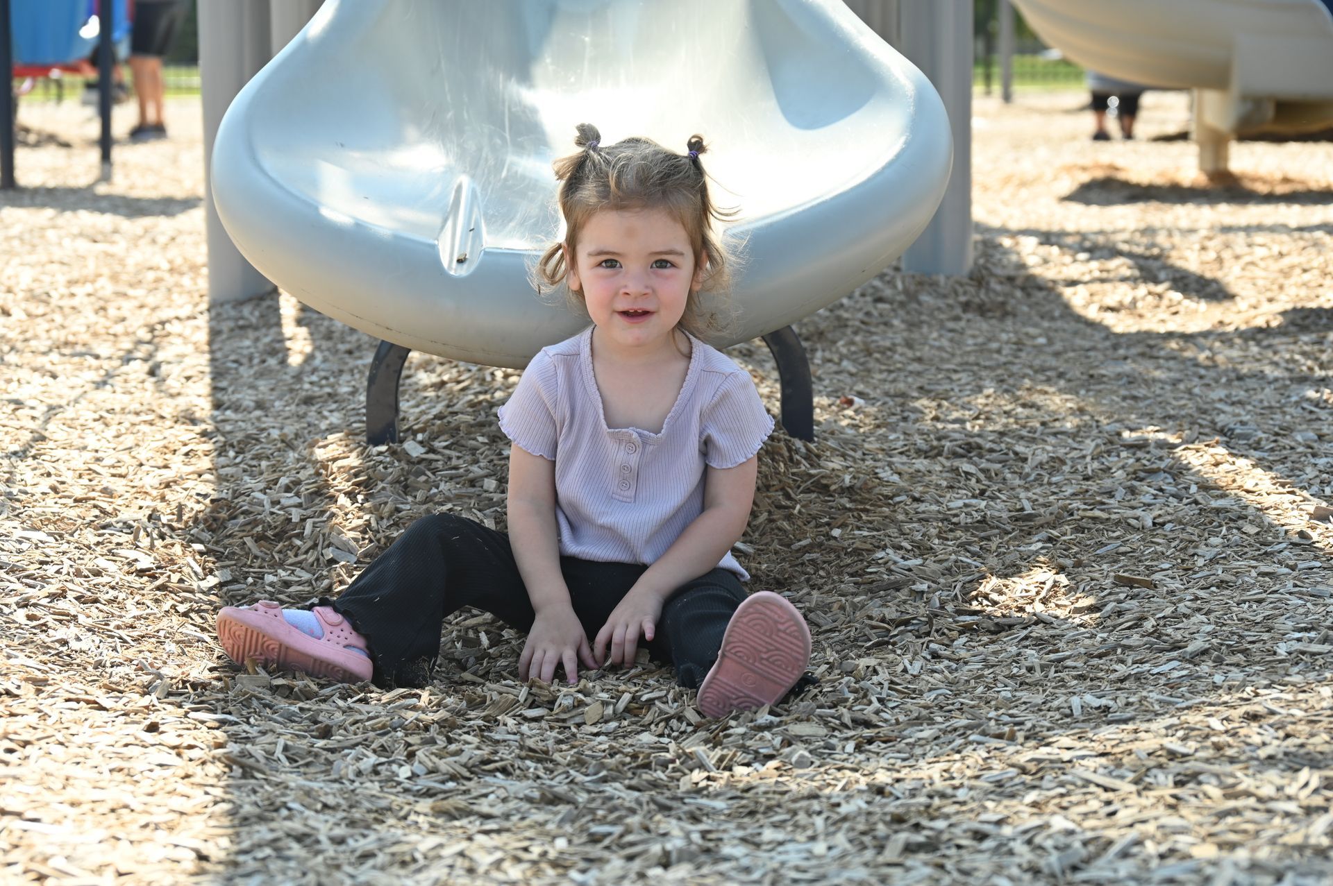 Child sitting at the base of a slide at a playground, wearing light purple shirt, pink shoes.