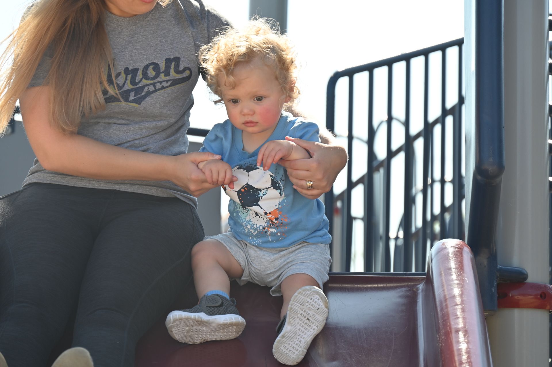 Woman helping a toddler on a playground slide. The child has curly red hair and is wearing a blue shirt.